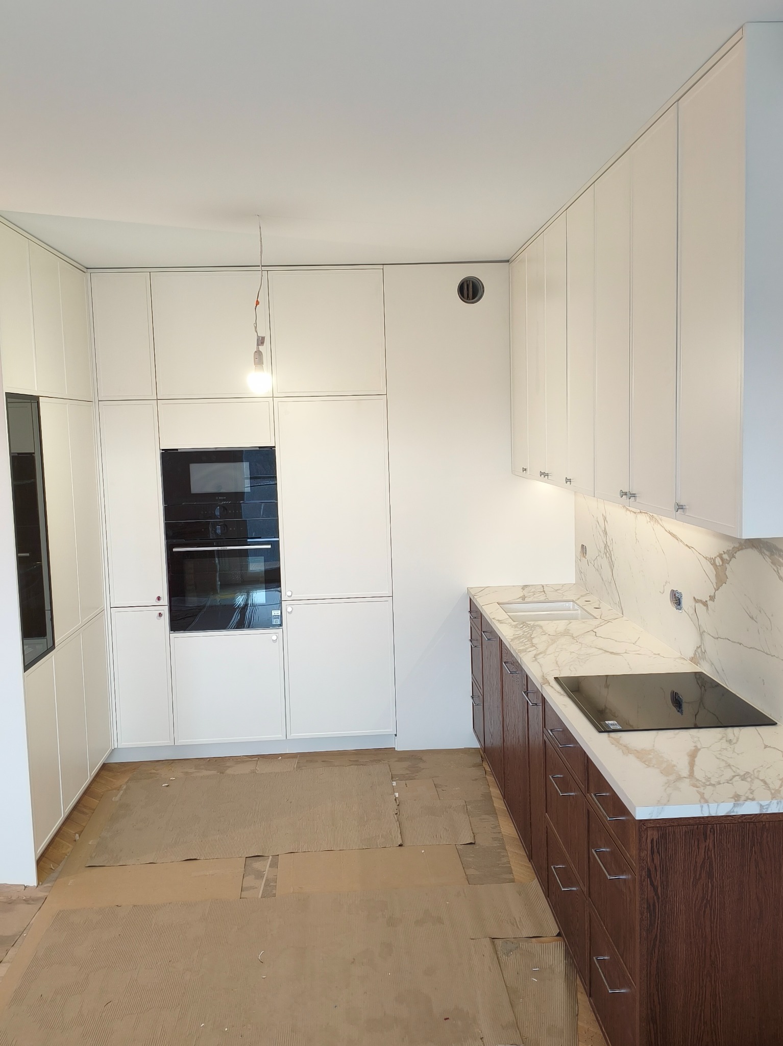Biała zabudowa kuchenna z drewnianymi szafkami dolnymi i marmurowym blatem w Bratkowicach. Widoczny piekarnik i płyta indukcyjna. Podłoga zabezpieczona kartonem.
