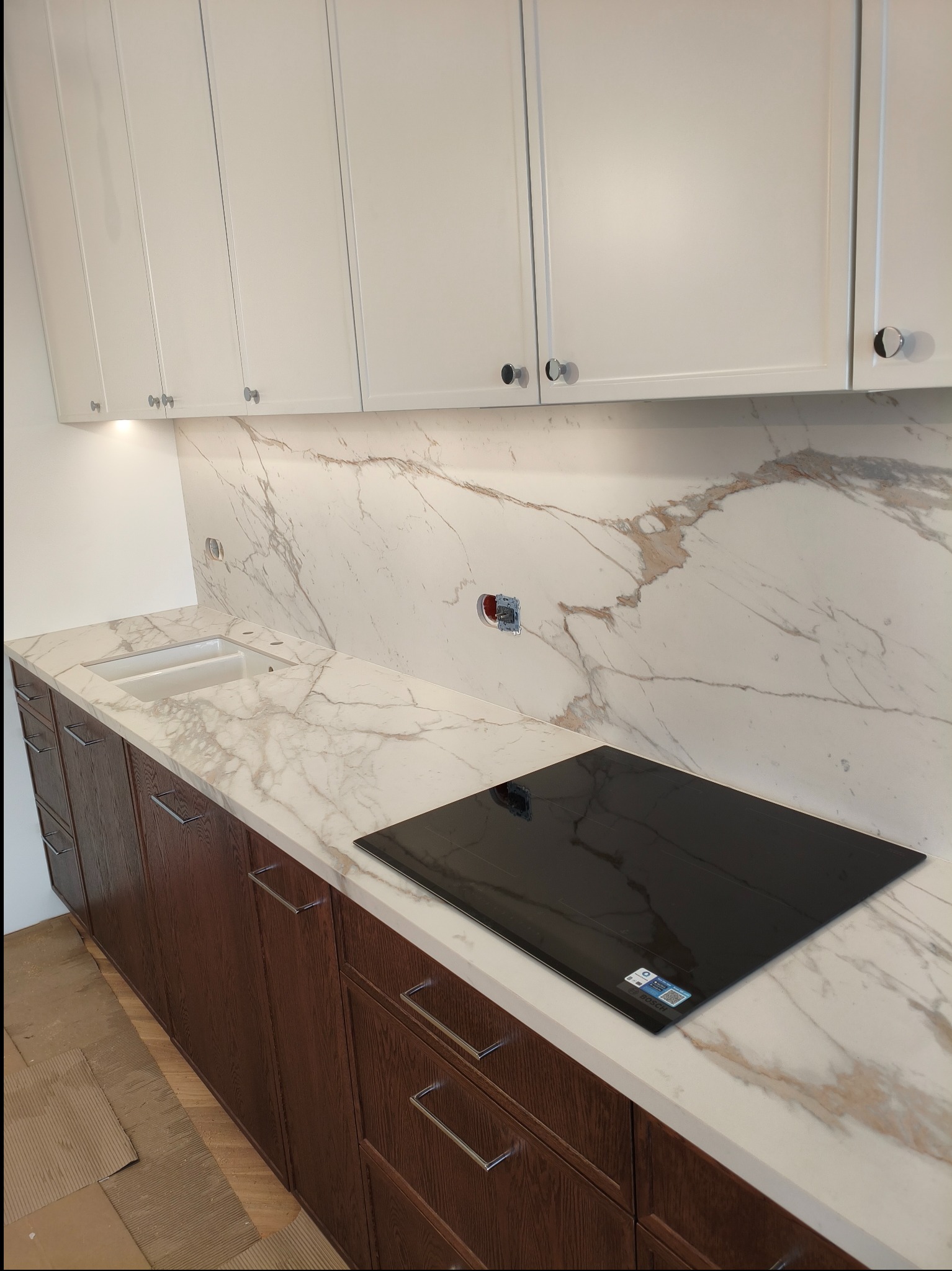 Nowoczesna kuchnia z białymi szafkami górnymi i ciemnobrązowymi dolnymi. Marmurowy blat i backsplash z wbudowaną płytą indukcyjną i zlewozmywakiem.