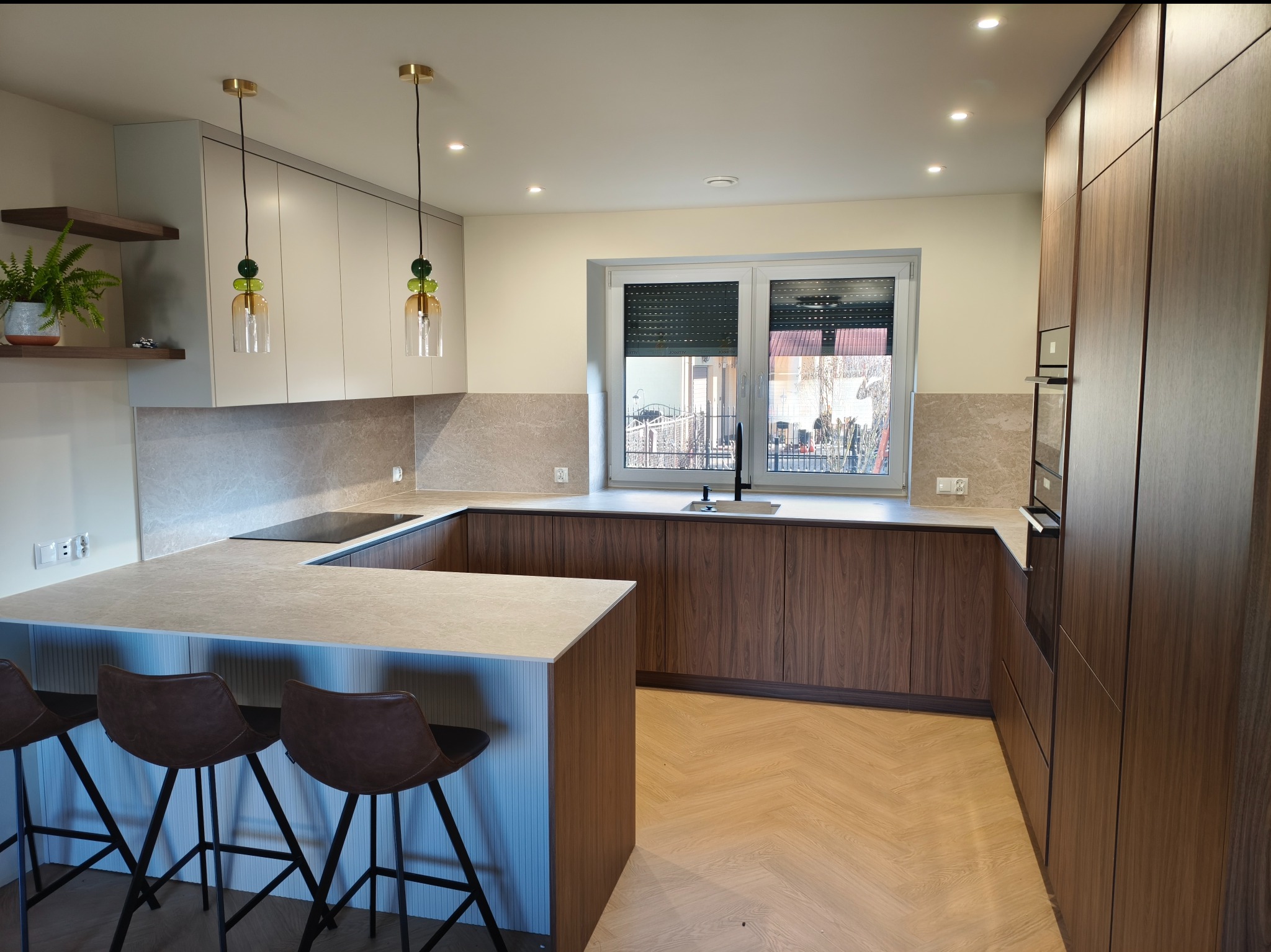 Nowoczesna kuchnia w kształcie litery U z drewnianymi frontami i blatem imitującym kamień, oświetlona wiszącymi lampami z zielonymi akcentami i trzema hokerami.