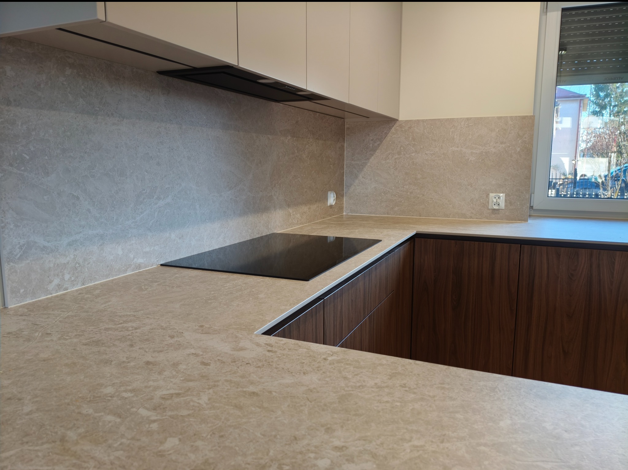 Nowoczesna kuchnia z blatem i ścianą w odcieniach beżu, z wbudowaną płytą indukcyjną i ciemnymi szafkami dolnymi. Minimalistyczny design, okno z żaluzjami w tle.