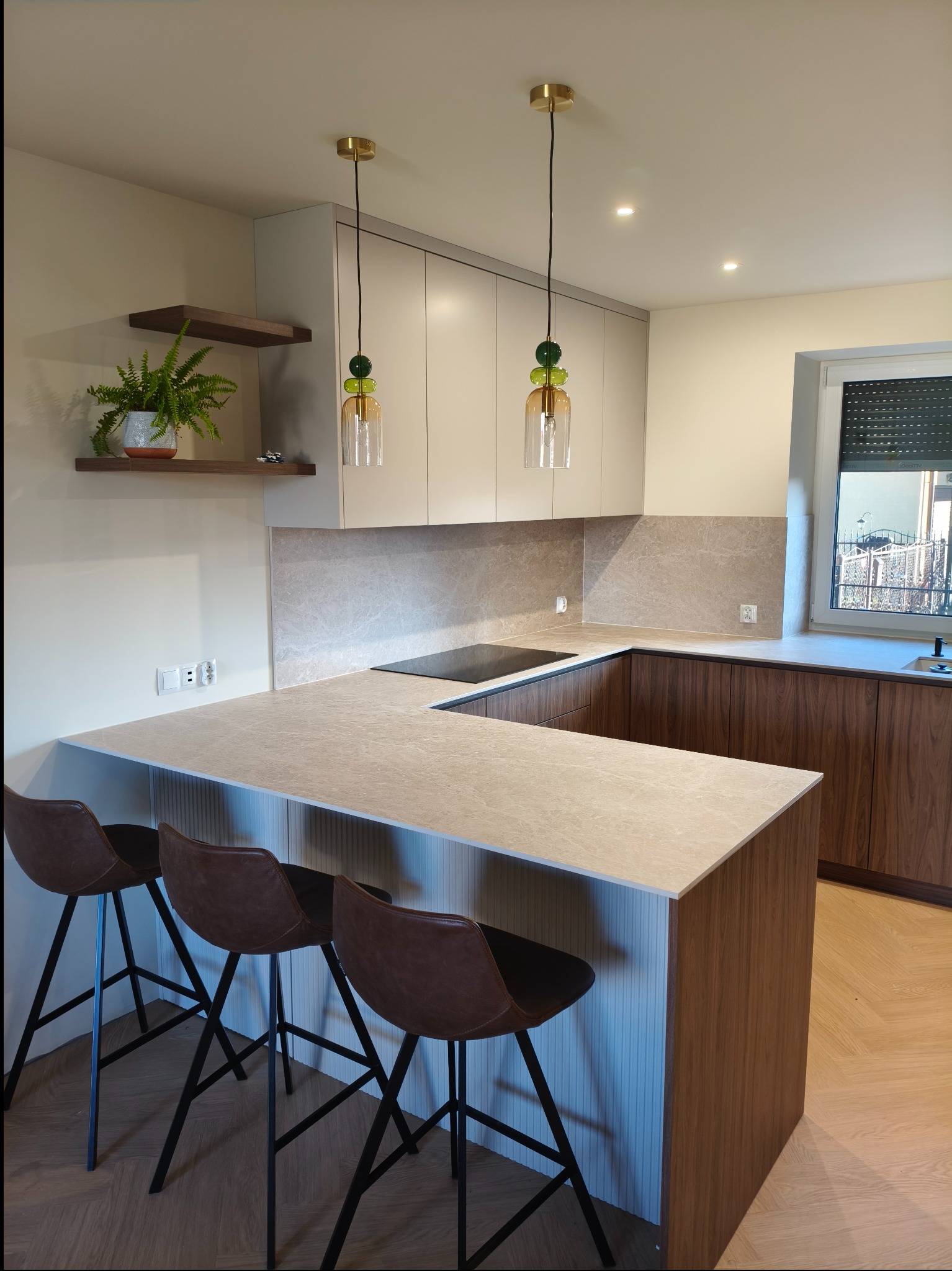 Nowoczesna kuchnia z wyspą i hokerami. Jasne szafki górne, drewniane dolne. Blat z kamienia. Nad wyspą wiszą lampy z kolorowego szkła. Na ścianie półka z paprotką.