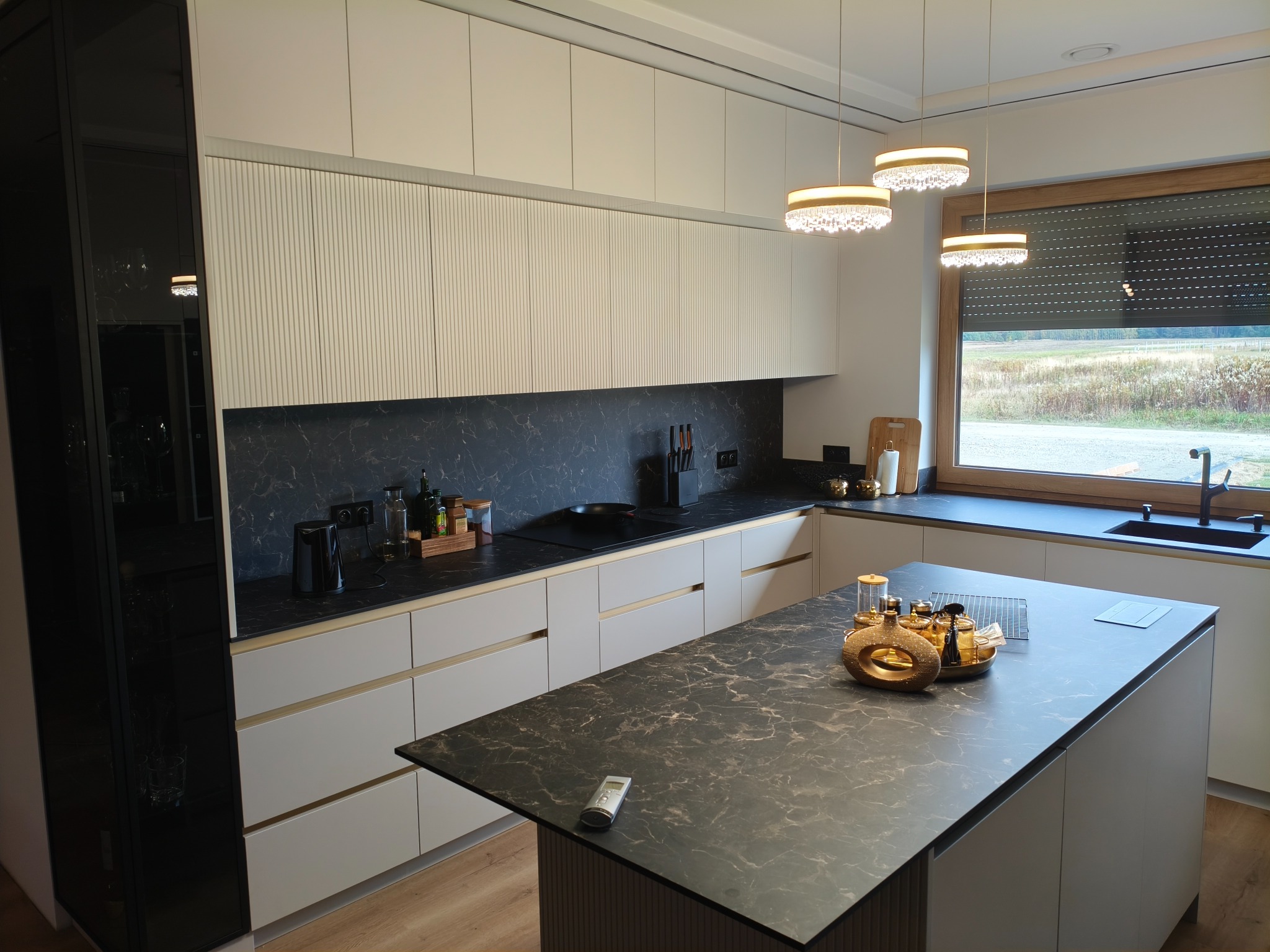 Nowoczesna kuchnia z wyspą, fronty szafek w odcieniach bieli i szarości, blat imitujący marmur. Nad wyspą dekoracyjne lampy. Widok na zewnątrz przez okno.