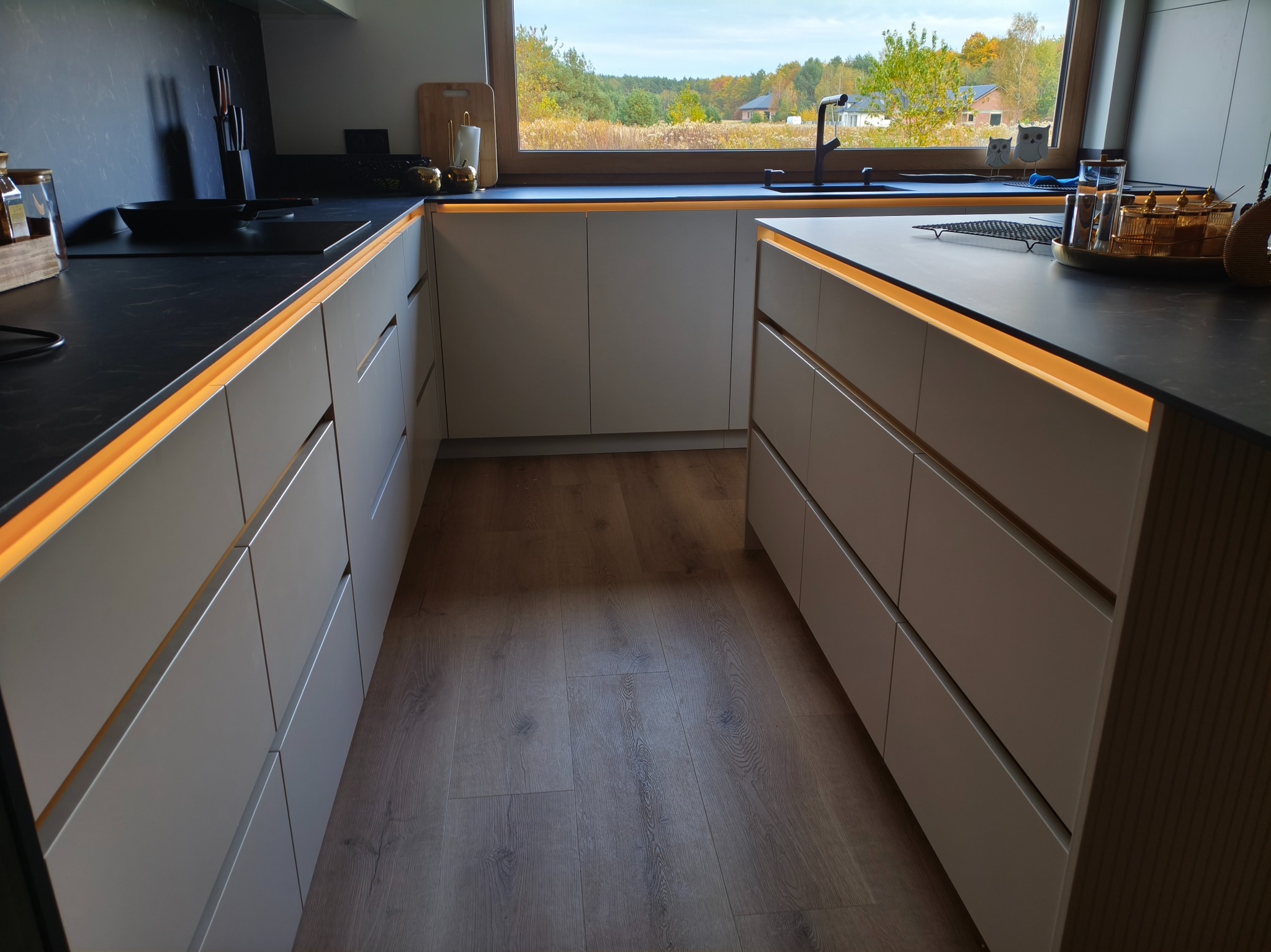 Nowoczesna kuchnia z szarymi szafkami i czarnym blatem, oświetlona listwami LED. Widok na okno z zielenią w tle. Minimalistyczny design, drewniana podłoga.