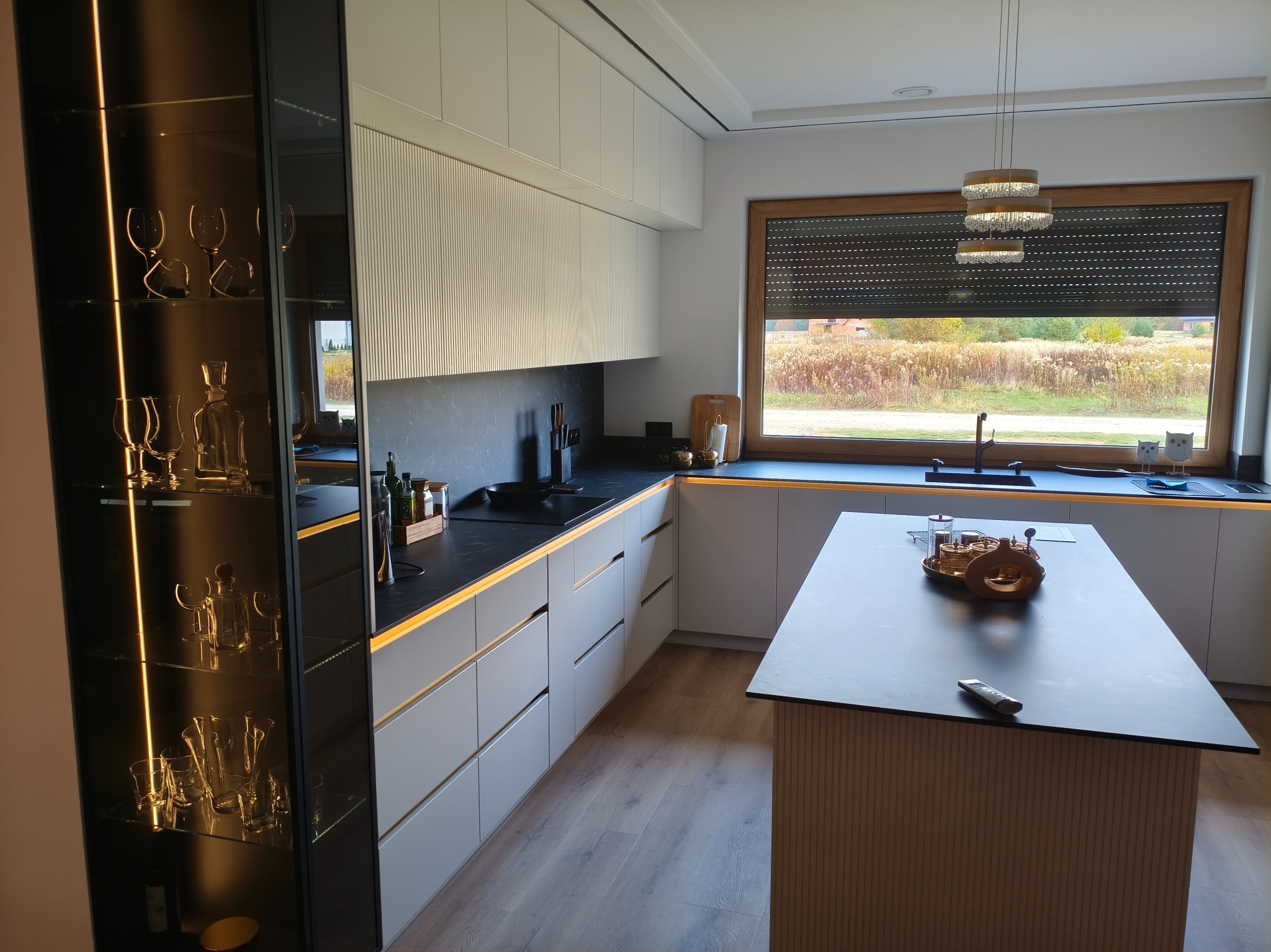 Nowoczesna kuchnia z szarymi szafkami, czarnym blatem i oświetleniem LED. Na wyspie kuchennej dekoracyjny zestaw. Za oknem widok na zewnątrz.