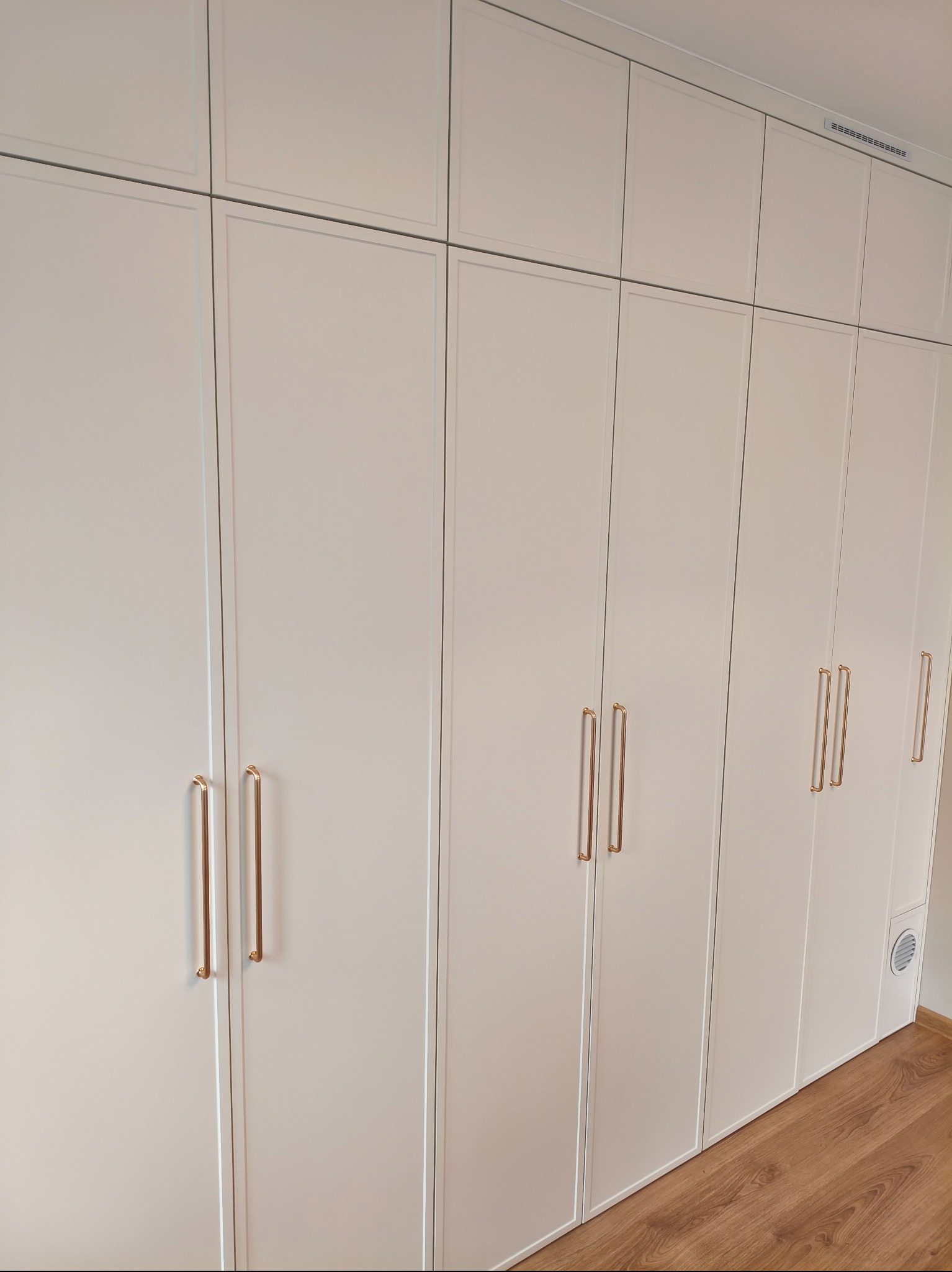 Biała szafa wnękowa z frezowanymi frontami i złotymi uchwytami, sięgająca sufitu, z widoczną kratką wentylacyjną w dolnej części i drewnianą podłogą.