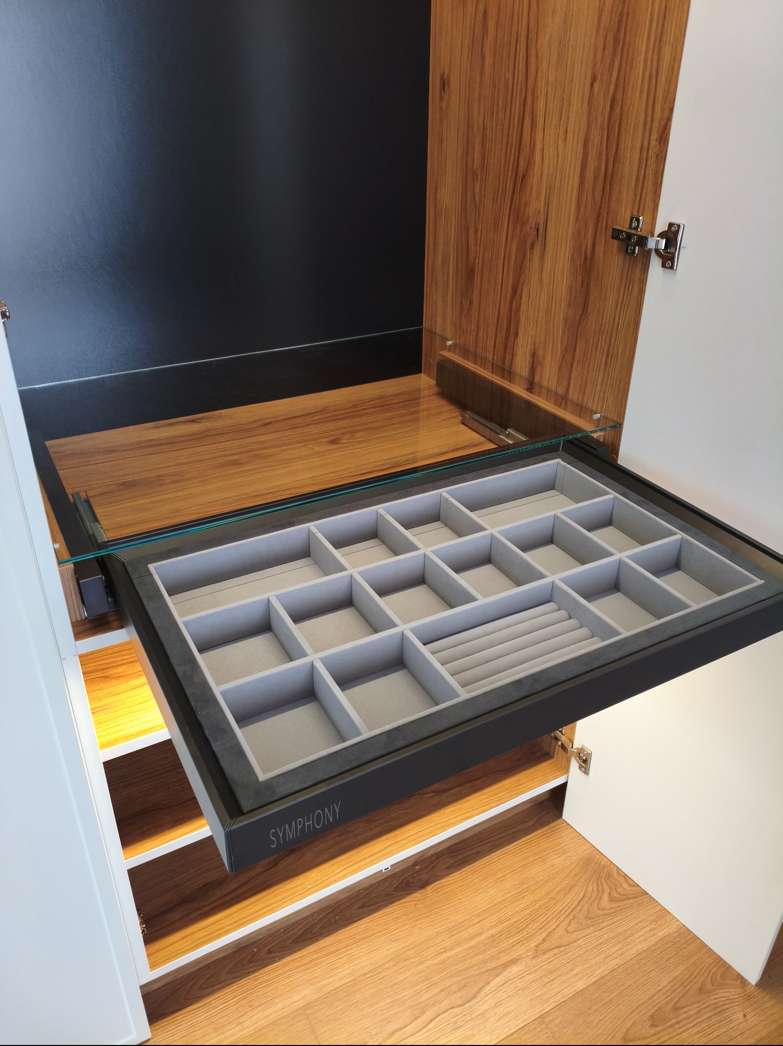 Wysuwana szuflada z przegródkami na biżuterię w szafie wnękowej. Szuflada z czarną ramą i szarym wnętrzem, widoczne drewniane elementy szafy i podłoga.