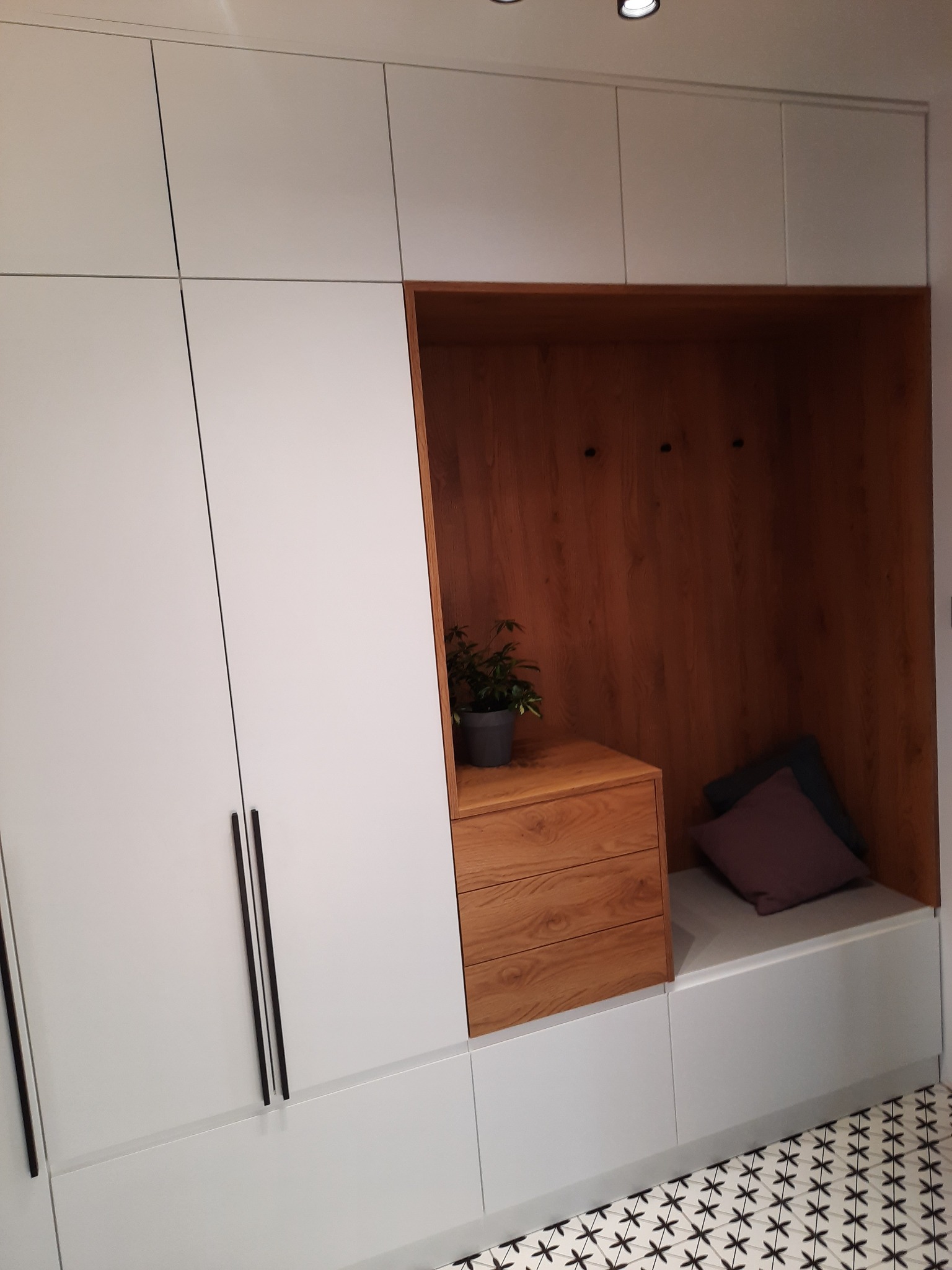 Biała szafa w przedpokoju z drewnianą wnęką z siedziskiem, drewnianą komodą i wieszakami, na komodzie stoi doniczka z rośliną, na siedzisku leżą poduszki, na podłodze wzorzyste płytki.