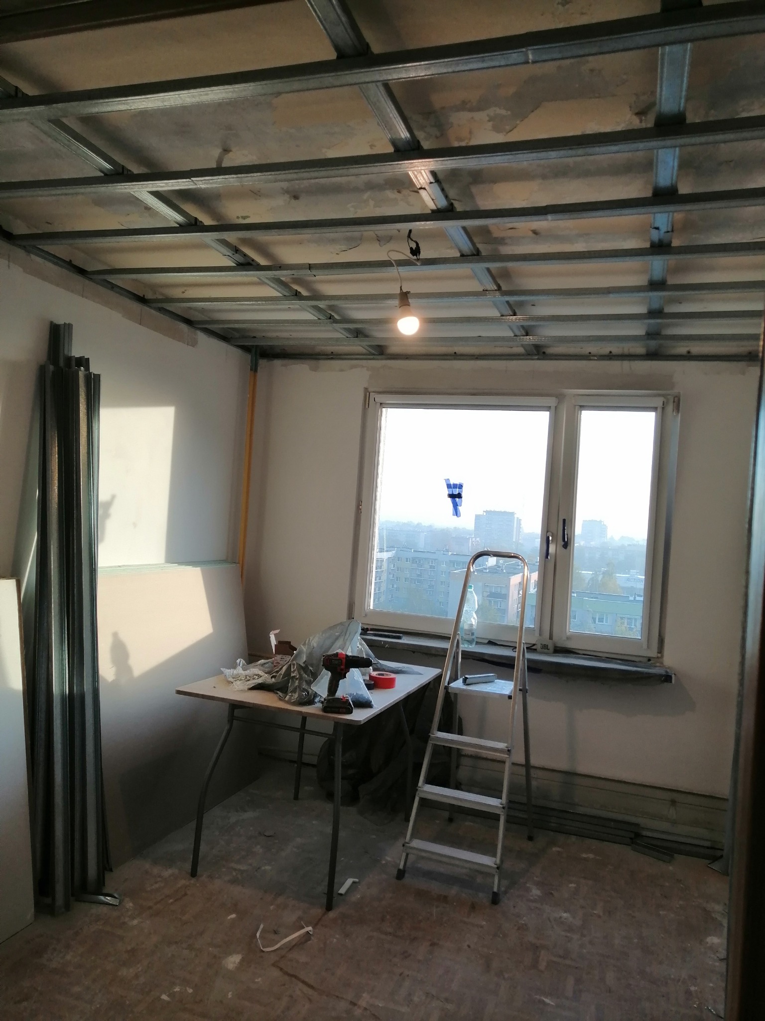 Montaż metalowej konstrukcji sufitu podwieszanego z widocznymi profilami, narzędziami na stole i stosem płyt kartonowo-gipsowych opartych o ścianę w pokoju z oknem.
