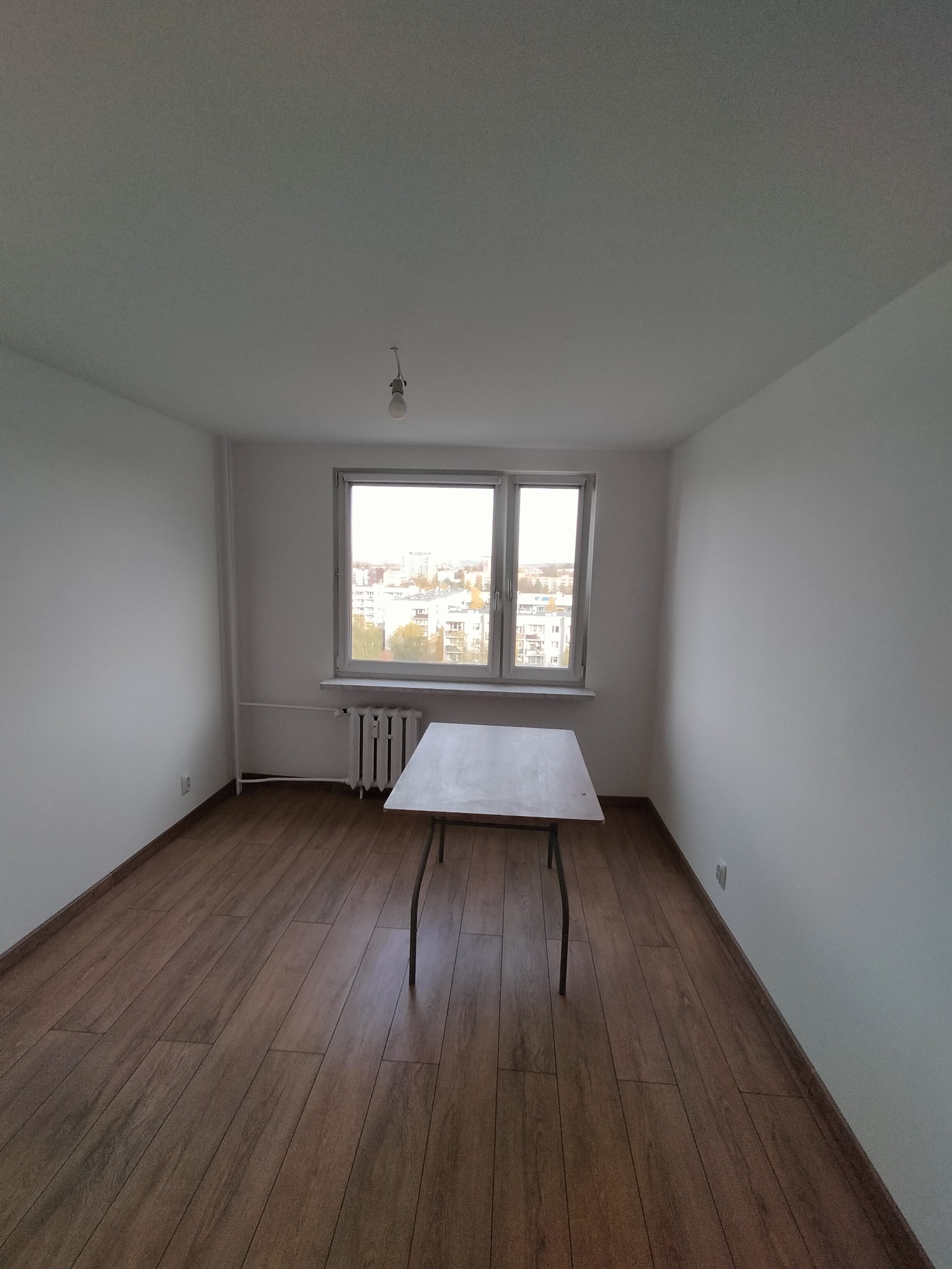 Pusty pokój po remoncie z panelami podłogowymi, białymi ścianami i stołem na środku, widok z okna na miasto.