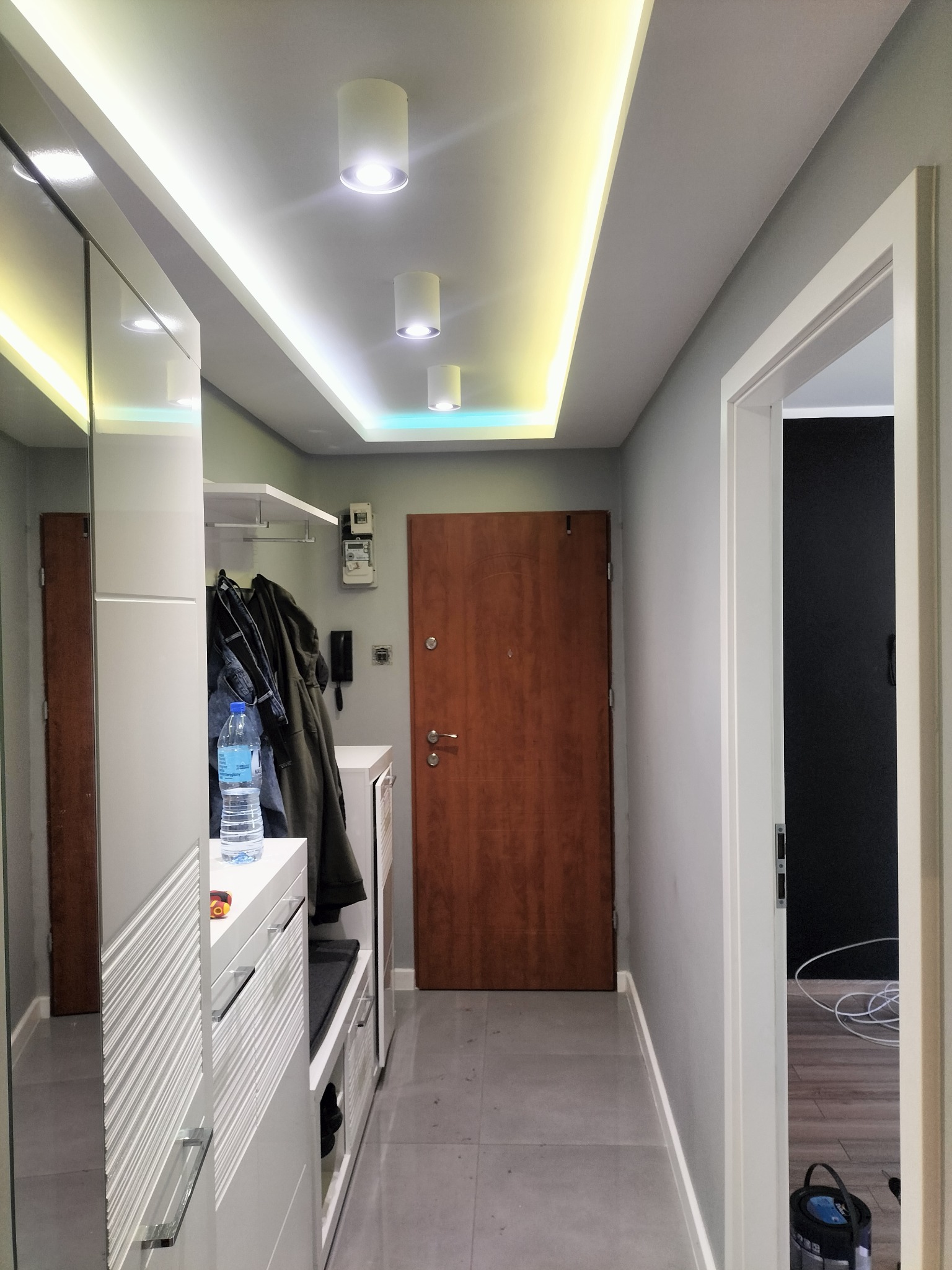 Nowoczesny przedpokój z oświetleniem LED w suficie podwieszanym, szafa z lustrem, w tle drewniane drzwi wejściowe i widok na pokój z ciemną ścianą.