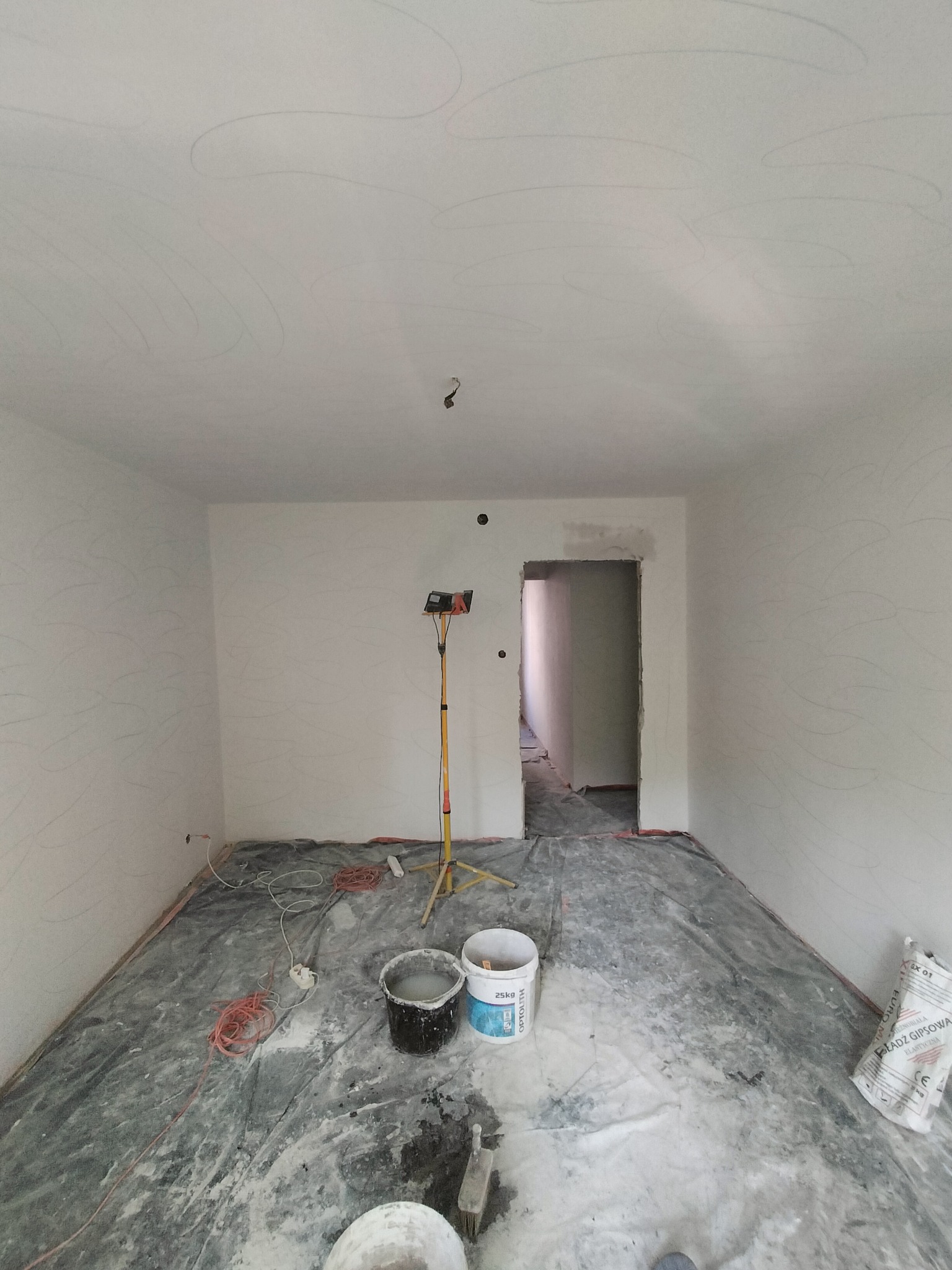 Pokój w trakcie przygotowania do malowania: ściany zagruntowane z widocznymi śladami ołówka, podłoga zabezpieczona folią, dwa wiadra z farbą, lampa robocza i worek z gładzią gipsową.