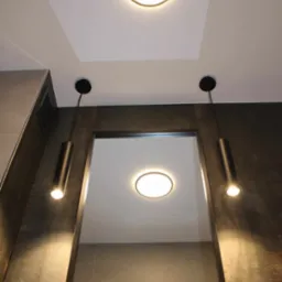 Nowoczesna łazienka z oświetleniem punktowym: dwa czarne, cylindryczne reflektory wiszące po bokach lustra oraz okrągła lampa sufitowa wpuszczana w podwieszany sufit, ciemna płytka imitująca metal...