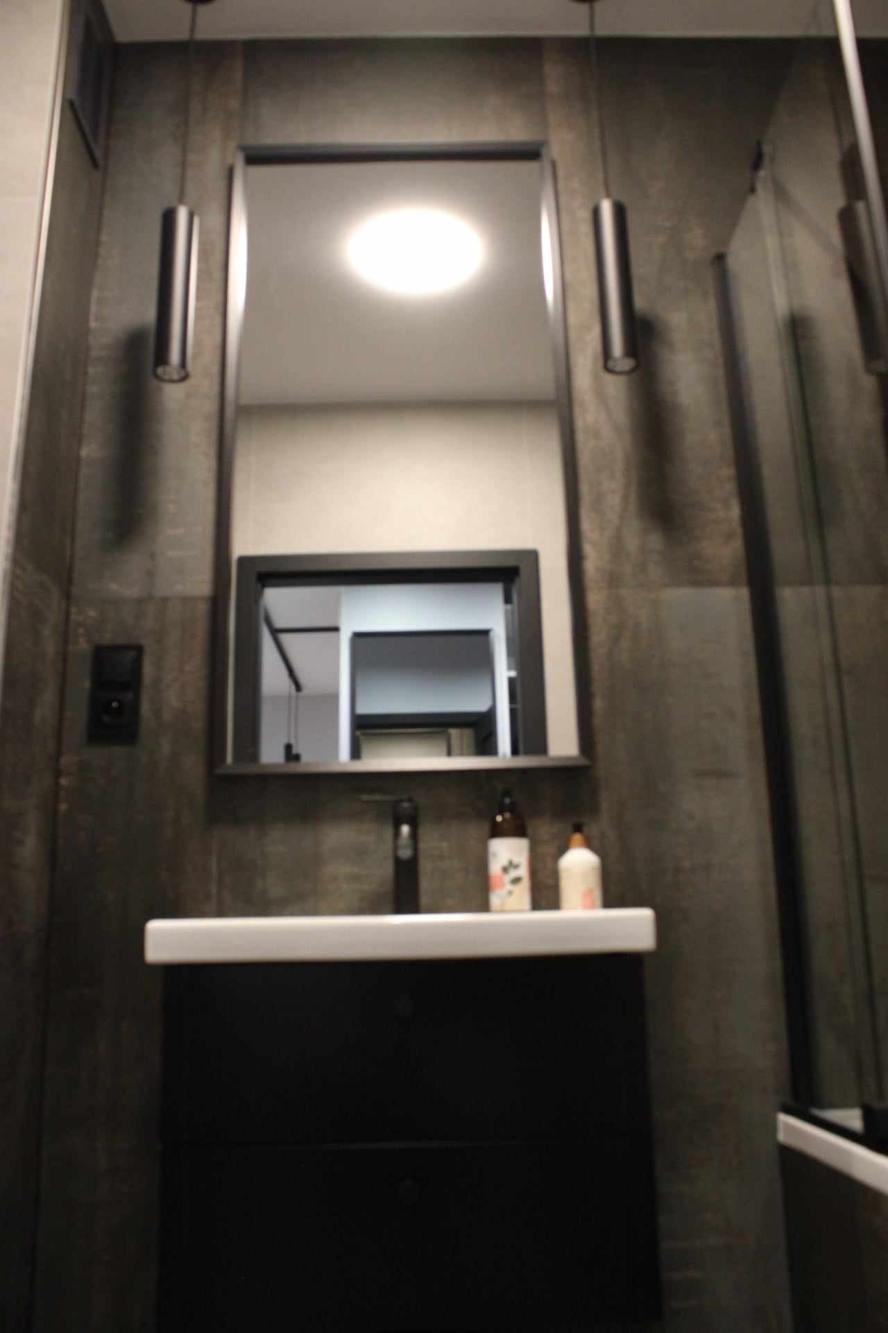 Nowoczesna łazienka z ciemnymi płytkami imitującymi kamień, białą umywalką nablatową na czarnej szafce, minimalistycznym lustrem w czarnej ramie i cylindrycznymi lampami wiszącymi.