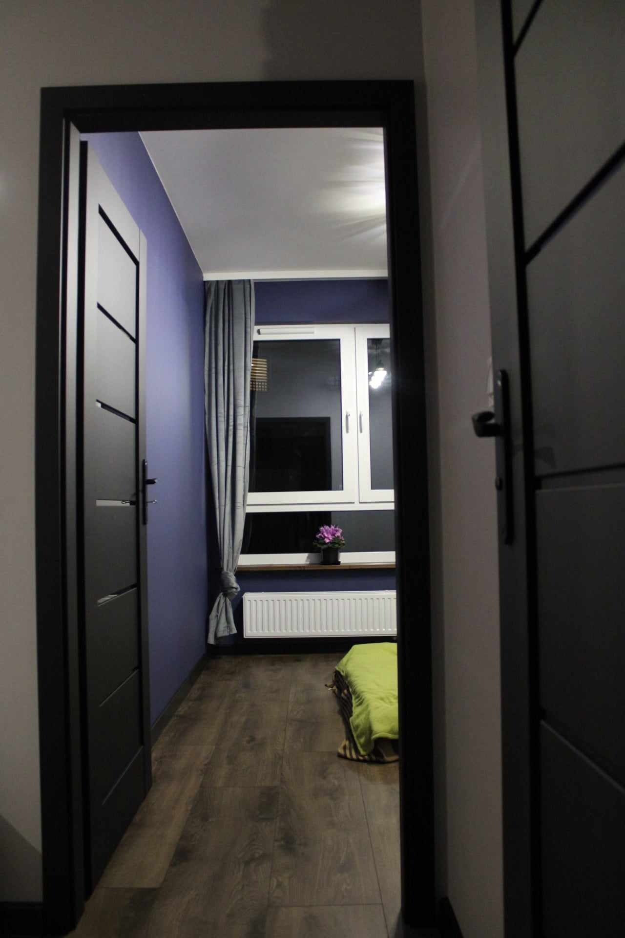 Wnętrze pokoju z ciemnymi panelami podłogowymi i fioletową ścianą, widoczne przez otwarte, czarne drzwi z dekoracyjnymi wstawkami, z oknem z szarą zasłoną i grzejnikiem pod nim oraz małym bukietem...