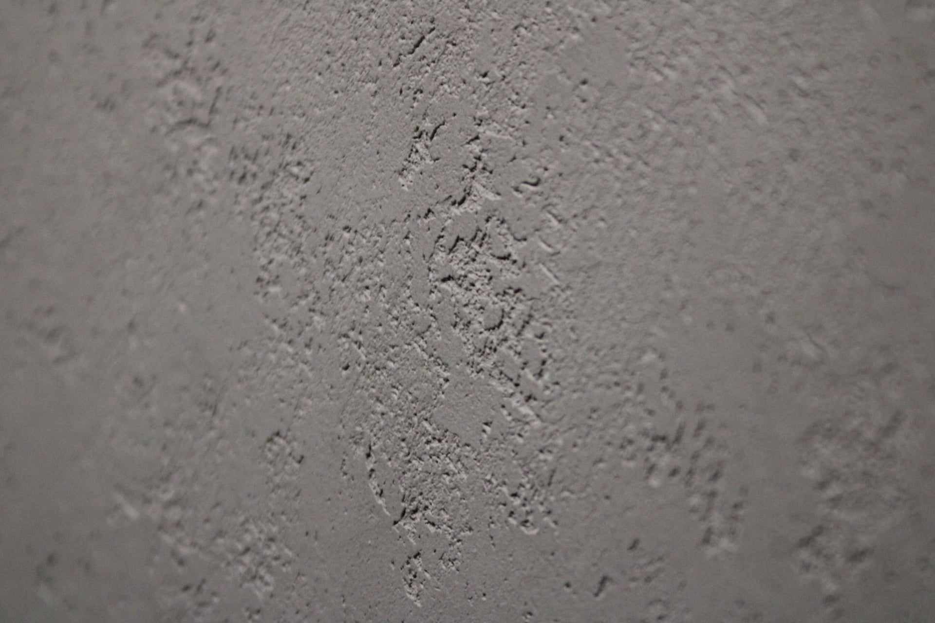 Szara ściana z wyraźną, nieregularną fakturą, oświetlona z boku, ukazująca gruboziarnistą strukturę tynku dekoracyjnego.