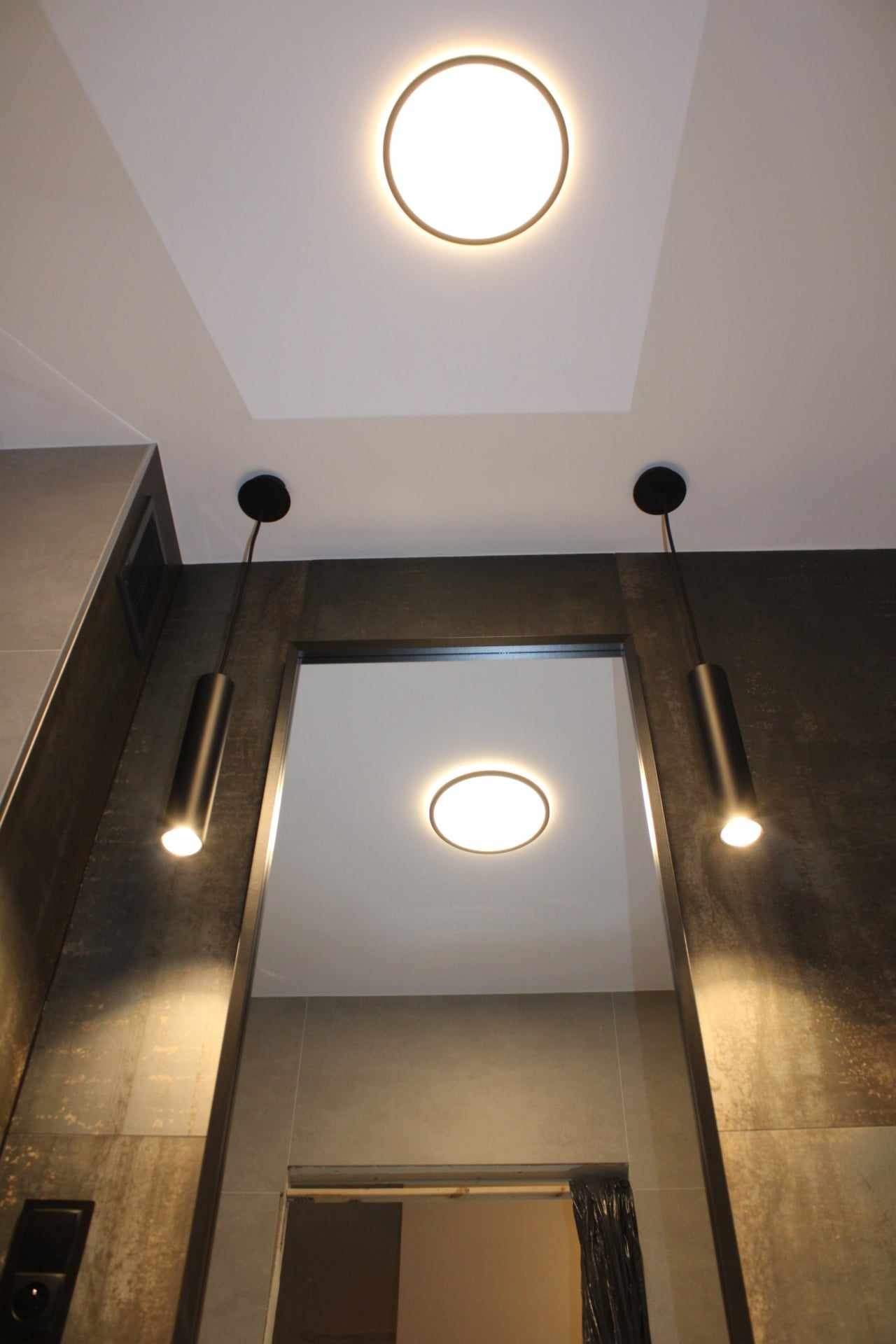 Nowoczesna łazienka z oświetleniem punktowym: dwa czarne, cylindryczne reflektory wiszące po bokach lustra oraz okrągła lampa sufitowa wpuszczana w podwieszany sufit, ciemna płytka imitująca metal...