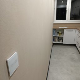 ARKADIUSZ ŻOCZEK - Jasna ściana z włącznikiem światła w nowocześnie wykończonej kuchni z szarymi płytkami podłogowymi i białymi szafkami. Widok na okno.