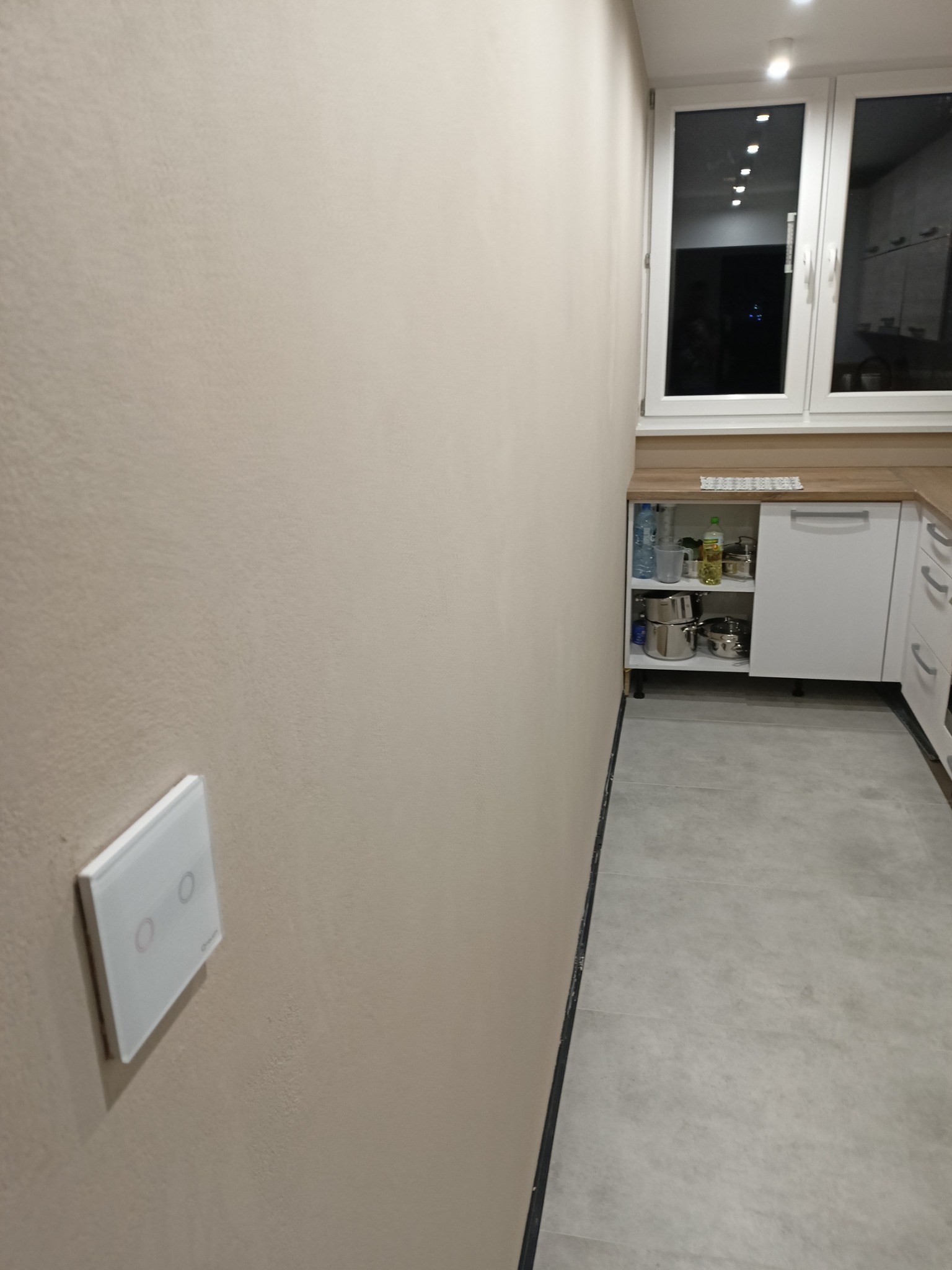 Jasna ściana z włącznikiem światła w nowocześnie wykończonej kuchni z szarymi płytkami podłogowymi i białymi szafkami. Widok na okno.