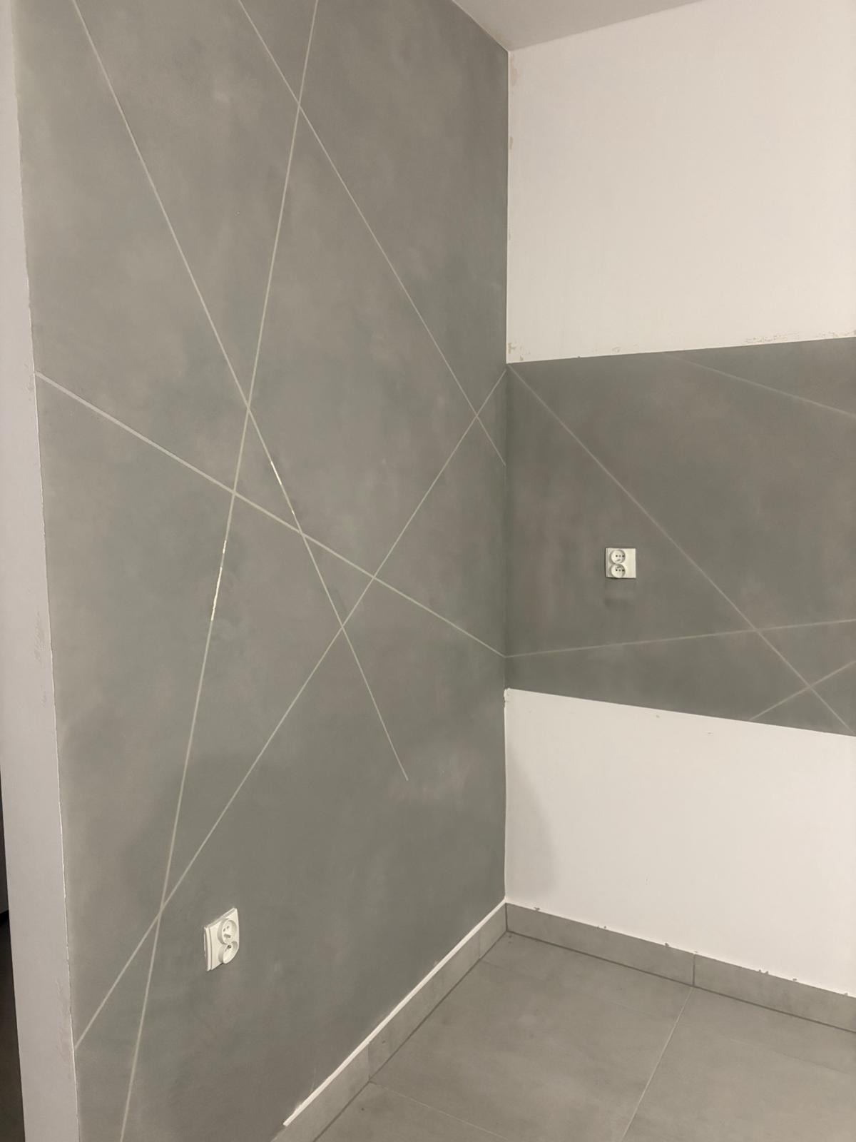 Szara ściana z dekoracyjnymi, jasnymi liniami tworzącymi geometryczny wzór. Widoczne gniazdka elektryczne i fragment podłogi z szarych płytek.