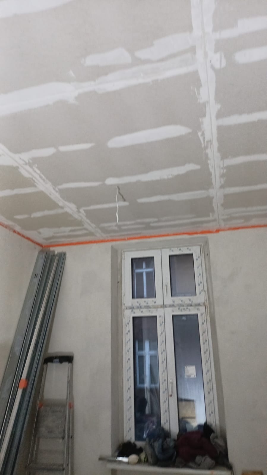 Sufit z płyt karton-gips, częściowo wyszpachlowany, widoczne profile, drabina i okno. Prace wykończeniowe w trakcie realizacji.