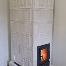 Biały piec kaflowy z paleniskiem, zdobiony złotymi uchwytami, stoi na podłodze z płytek w niebiesko-białe wzory, z widocznym fragmentem drewnianej podłogi.