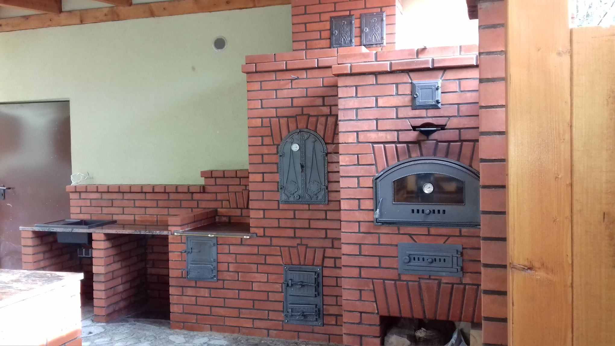Murowana kuchnia letnia z cegły z wbudowanym piecem chlebowym i blatem roboczym, ozdobne żeliwne drzwiczki piekarnika.