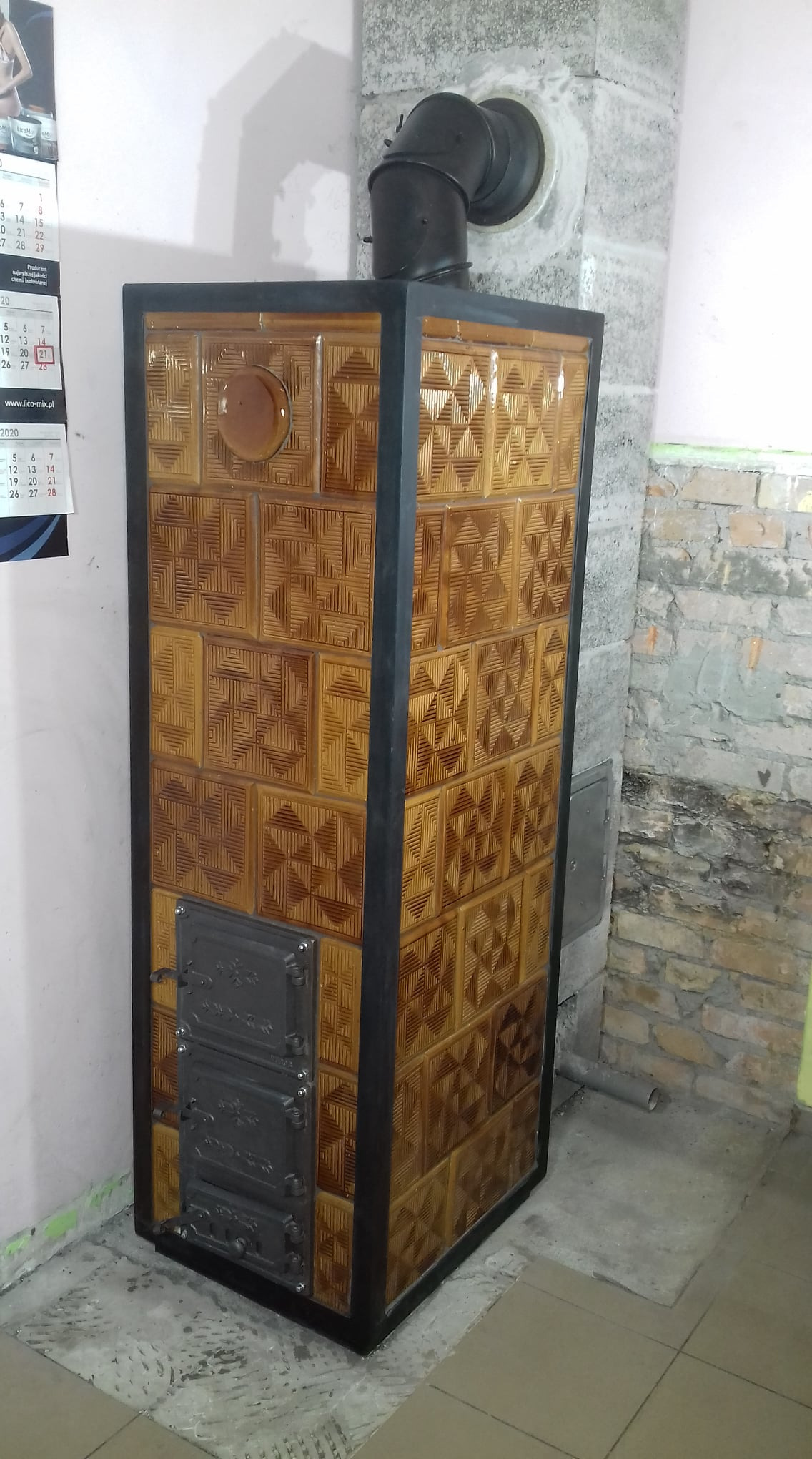 Kafelkowy piec z żeliwnymi drzwiczkami i czarną rurą odprowadzającą dym, widoczny na tle ściany z cegły i kalendarza.