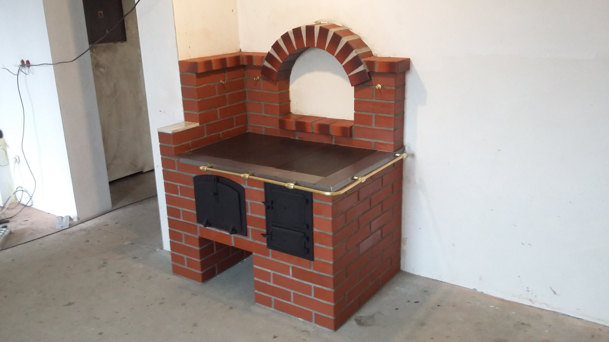 Murowany piec kuchenny z czerwonej cegły z czarnymi drzwiczkami, wykończony mosiężną rurką, z łukowym zwieńczeniem nad blatem.
