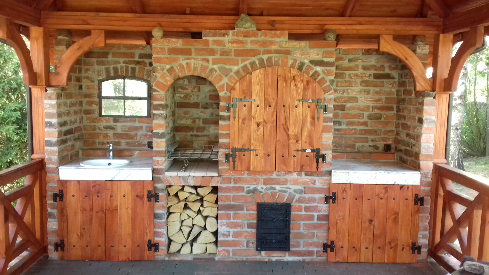 Murowana kuchnia letnia z cegły, wyposażona w zlew, grill z rusztem, schowek na drewno i zamykaną szafkę, całość pod drewnianą konstrukcją z widocznymi elementami dekoracyjnymi.