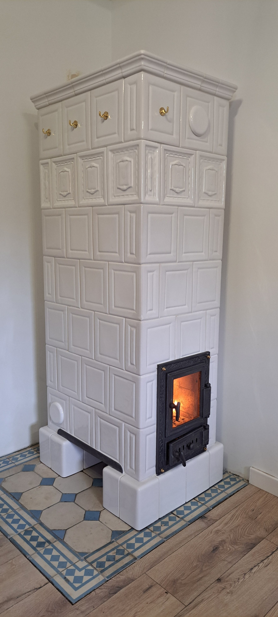 Biały piec kaflowy z paleniskiem, zdobiony złotymi uchwytami, stoi na podłodze z płytek w niebiesko-białe wzory, z widocznym fragmentem drewnianej podłogi.