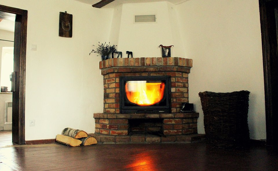 Otwarty kominek z cegły z płomieniami, ozdobiony figurkami i suszem na górze, obok kosz wiklinowy i drewno na podłodze.