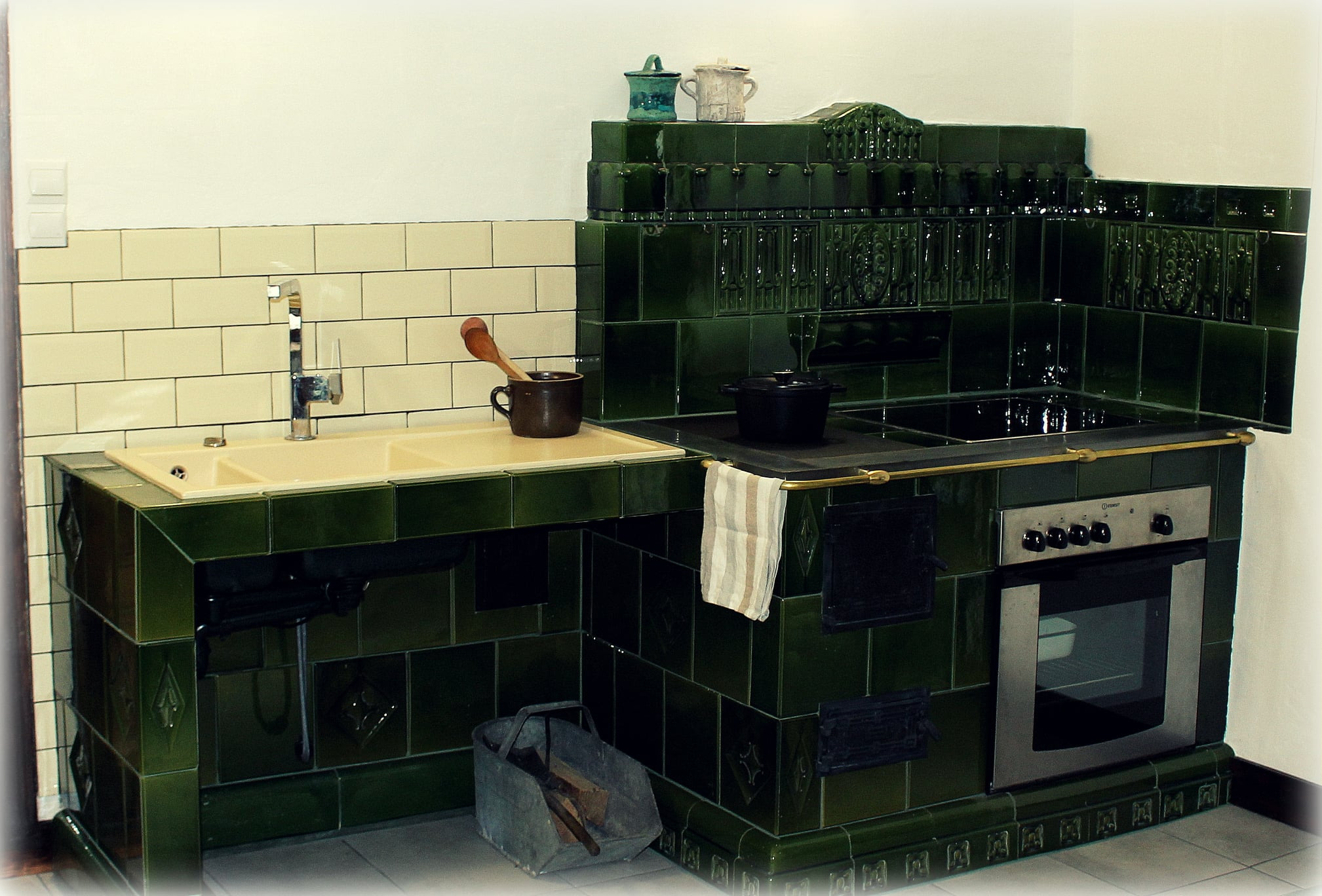 Kuchenna zabudowa z zielonych, ceramicznych kafli obejmująca zlew, płytę grzewczą i piekarnik, z drewnianą łyżką w kubku i metalowym koszem na drewno obok.