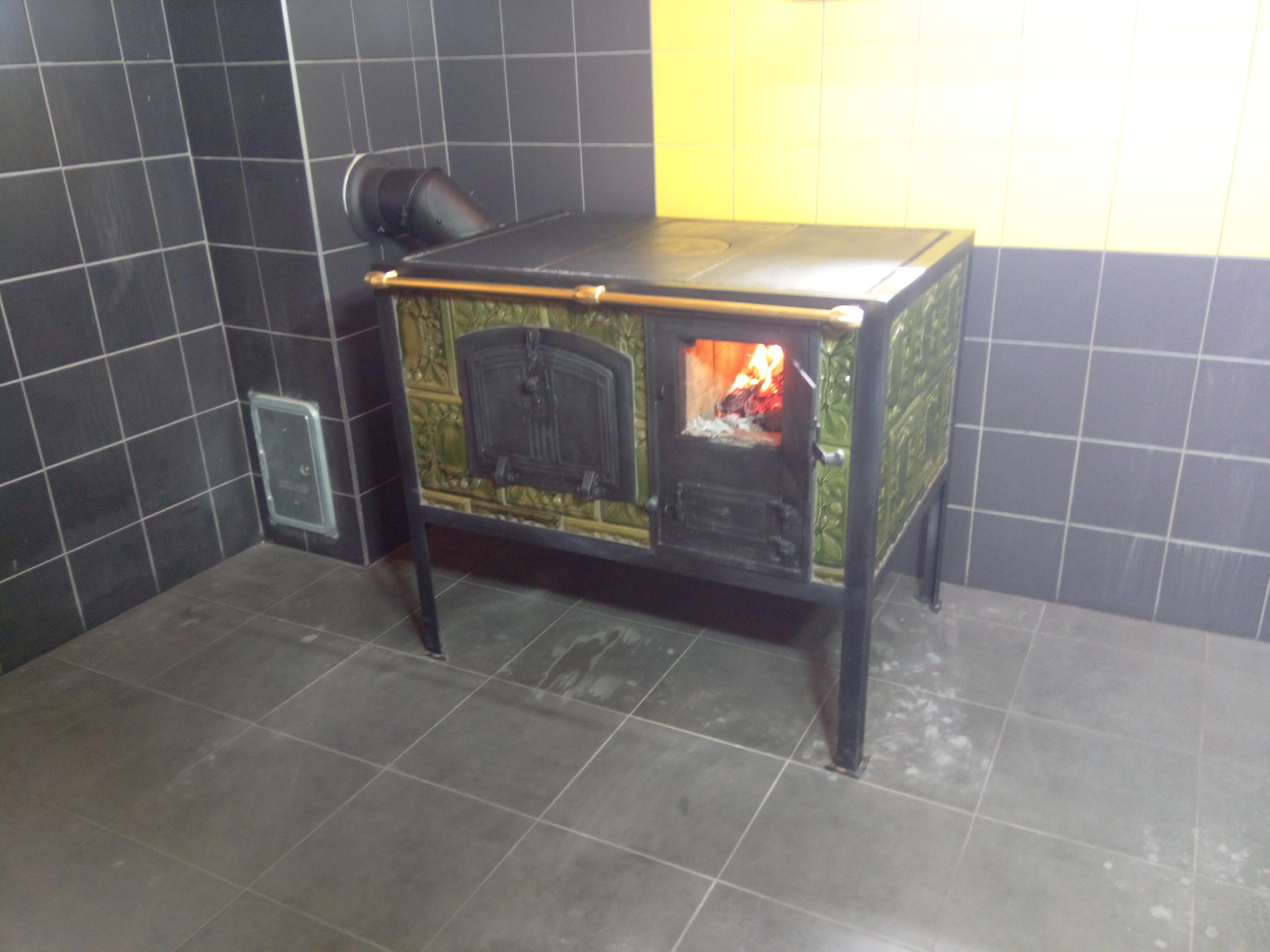 Kafelkowy piec kaflowy z palącym się drewnem w palenisku, w pomieszczeniu wyłożonym czarnymi i żółtymi płytkami ceramicznymi.