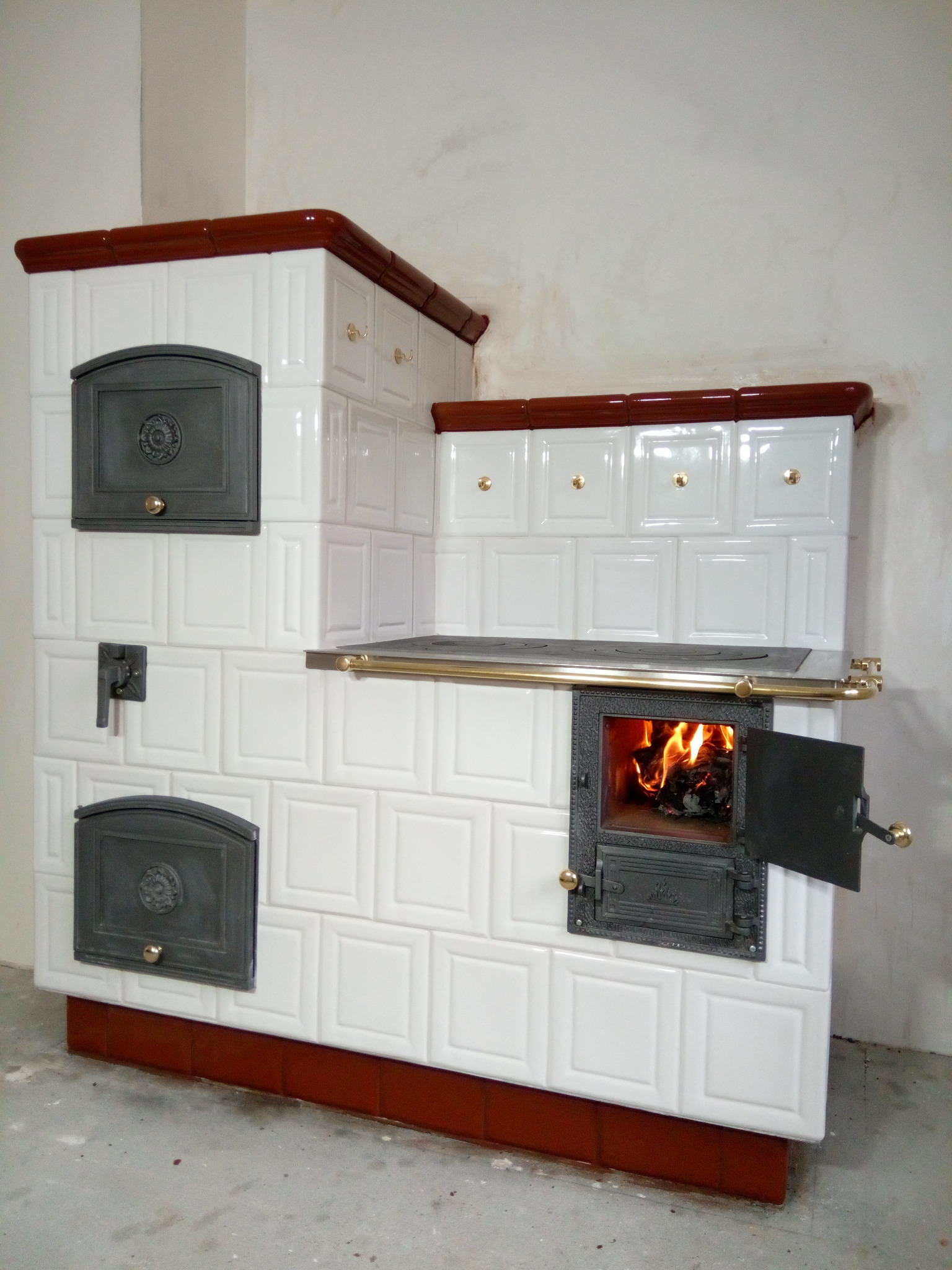 Biały piec kaflowy z brązowymi akcentami, otwarte drzwiczki paleniska ukazują płomienie, złote uchwyty i zawiasy.