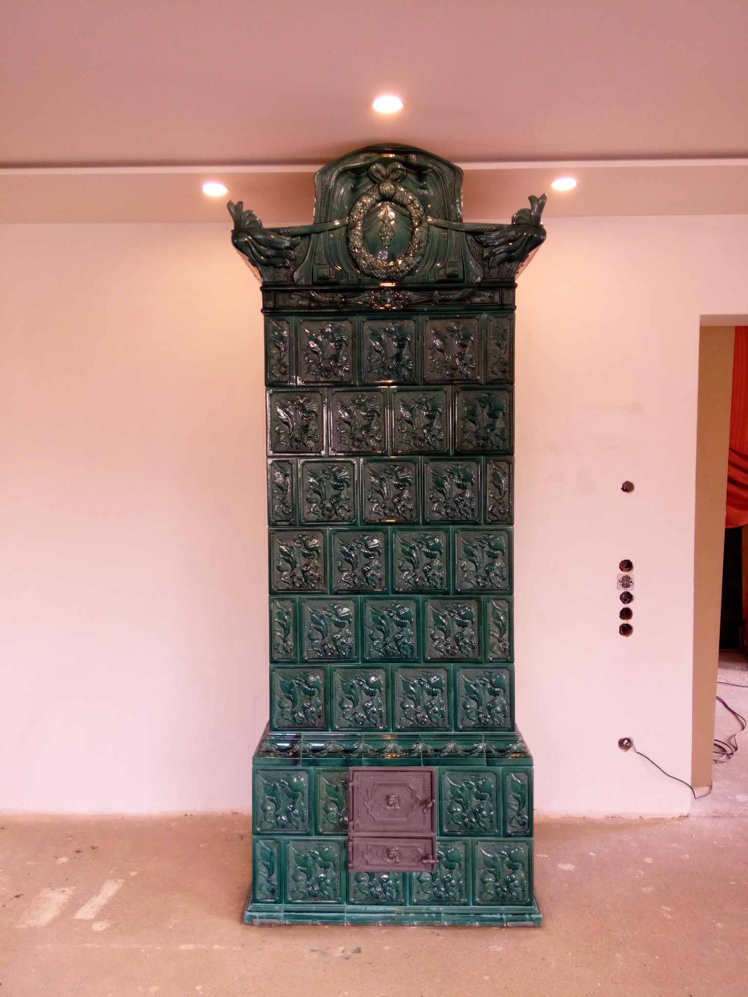 Wysoki, zielony piec kaflowy z ozdobnymi płaskorzeźbami kwiatowymi, zwieńczony dekoracyjną koroną, w surowym wnętrzu z widocznymi gniazdkami elektrycznymi.