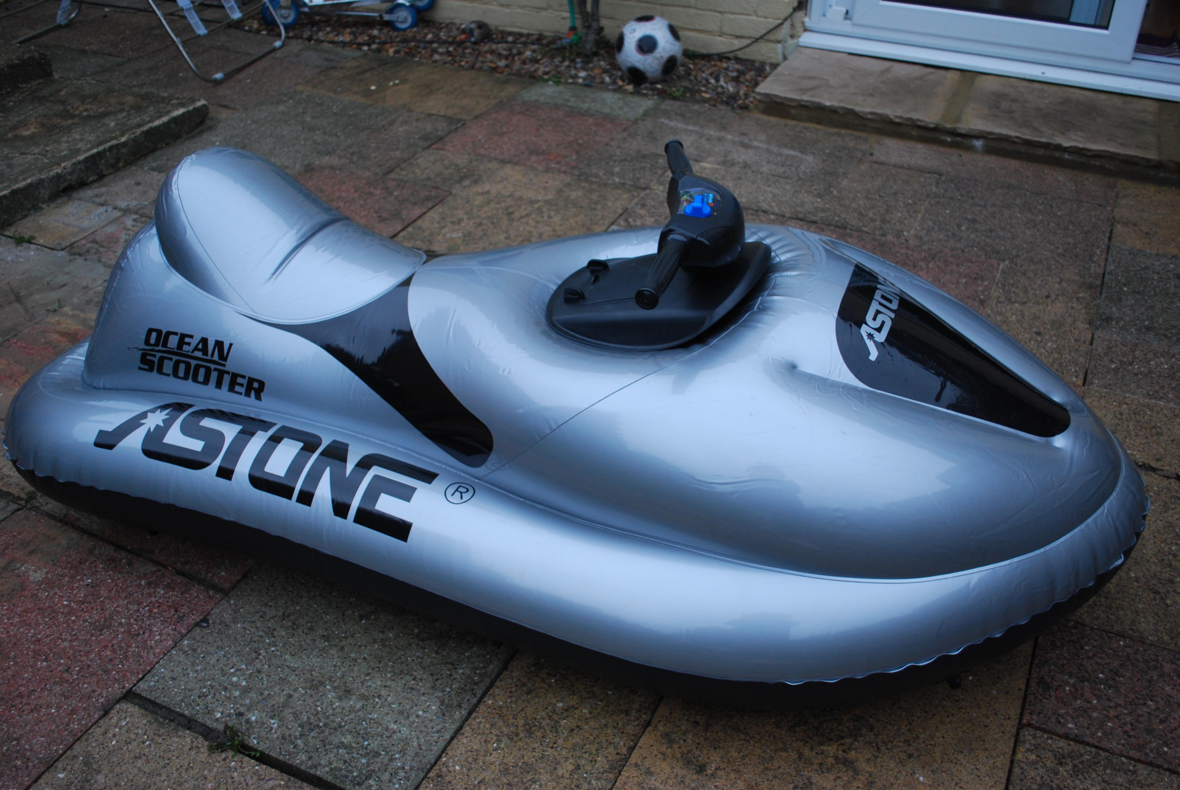 Niebiesko-szary, nadmuchiwany skuter wodny z czarnymi detalami i napisem 'Ocean Scooter' oraz 'ASTONE' leży na brukowanym podwórku z piłką do piłki nożnej i drzwiami w tle.