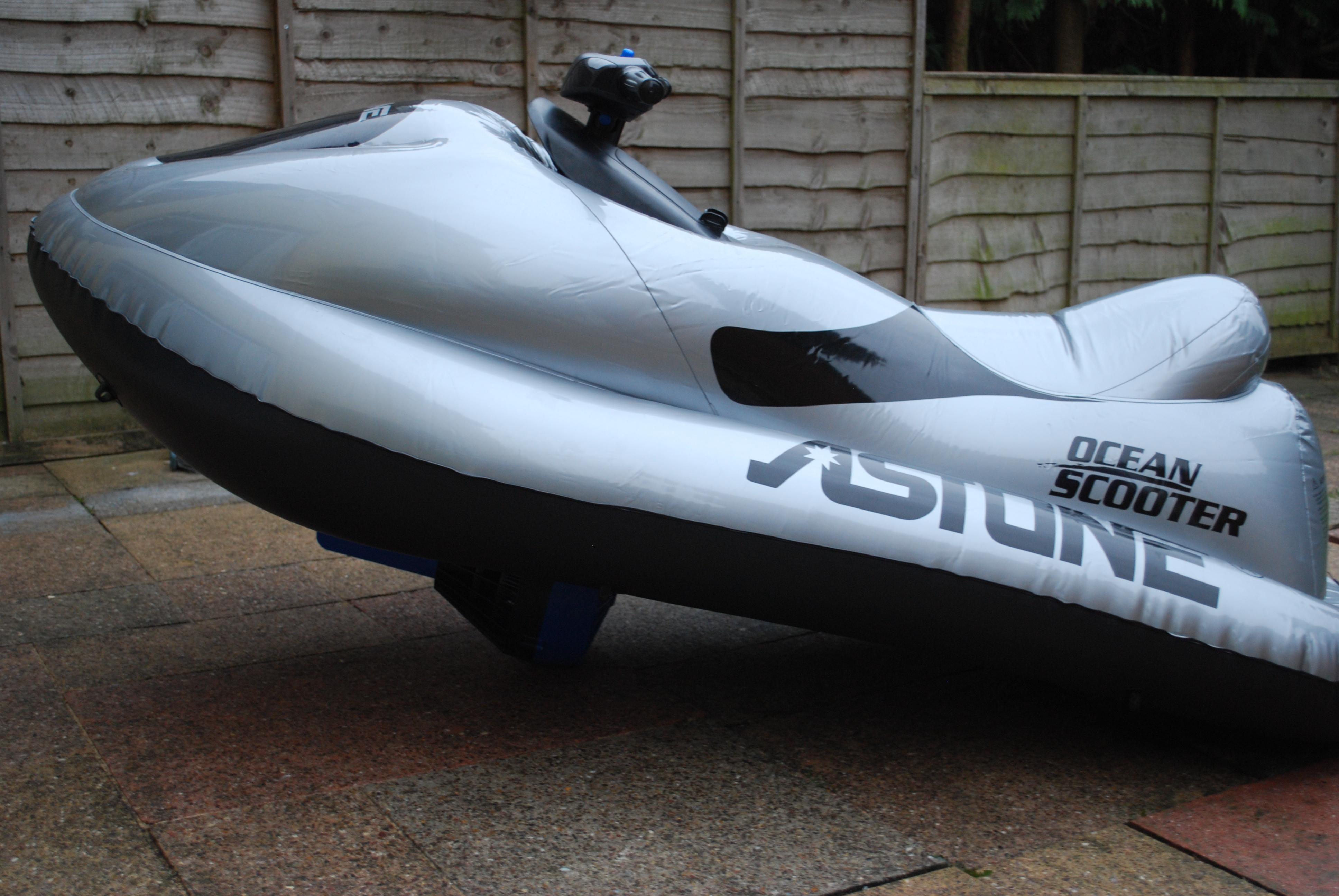 Szaro-czarny pompowany skuter wodny marki Astune Ocean Scooter stojący na brukowanej powierzchni, z drewnianym płotem w tle.