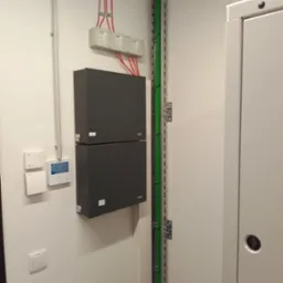 Dwa czarne moduły Roger zamontowane na ścianie obok przewodów w metalowej szynie i termostatu cyfrowego.