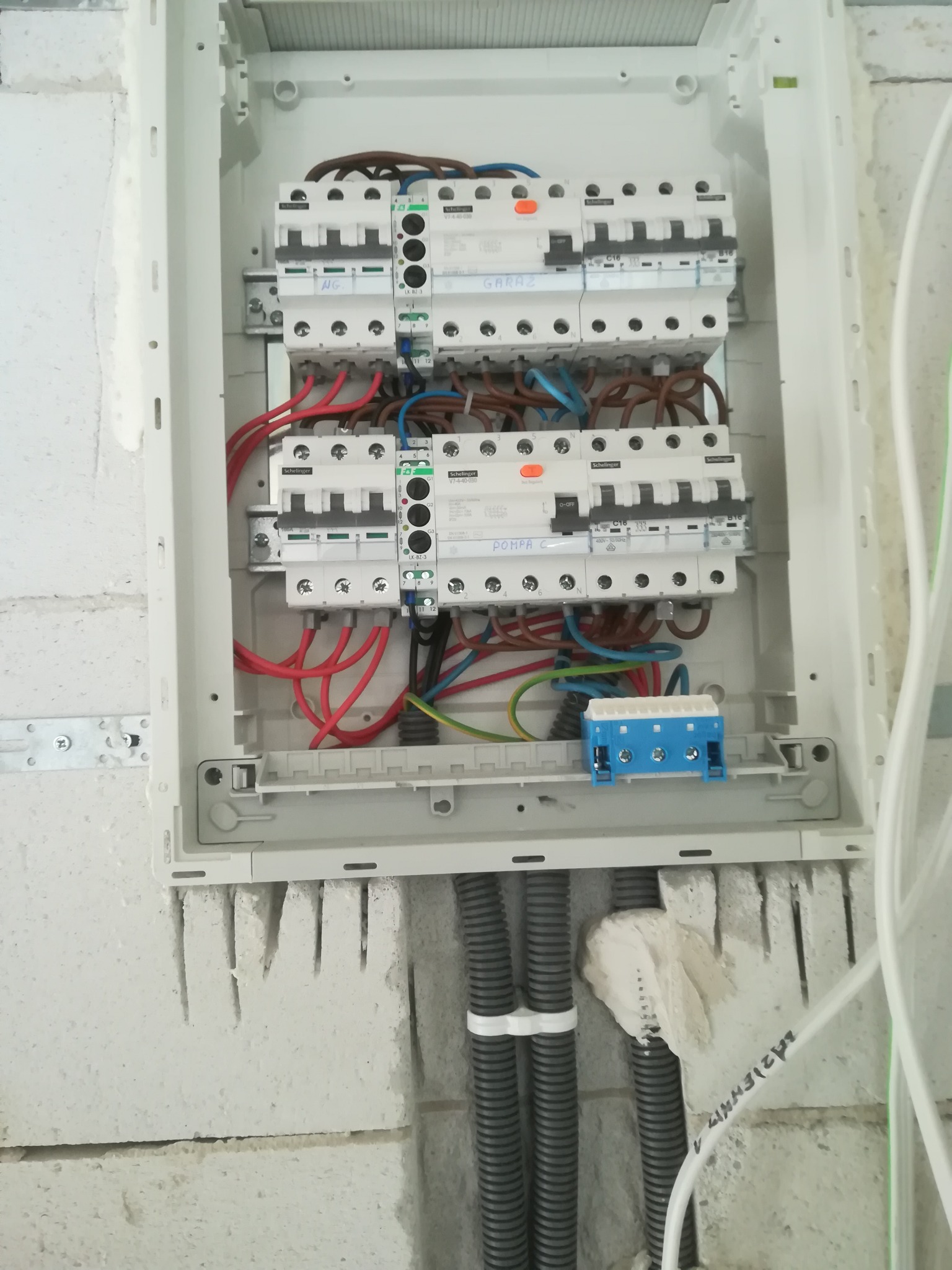 Otwarta skrzynka elektryczna z zamontowanymi bezpiecznikami Schilinger, widoczne kable w kolorach czerwonym, brązowym, niebieskim i zielono-żółtym, zamocowana na ścianie z betonu komórkowego...