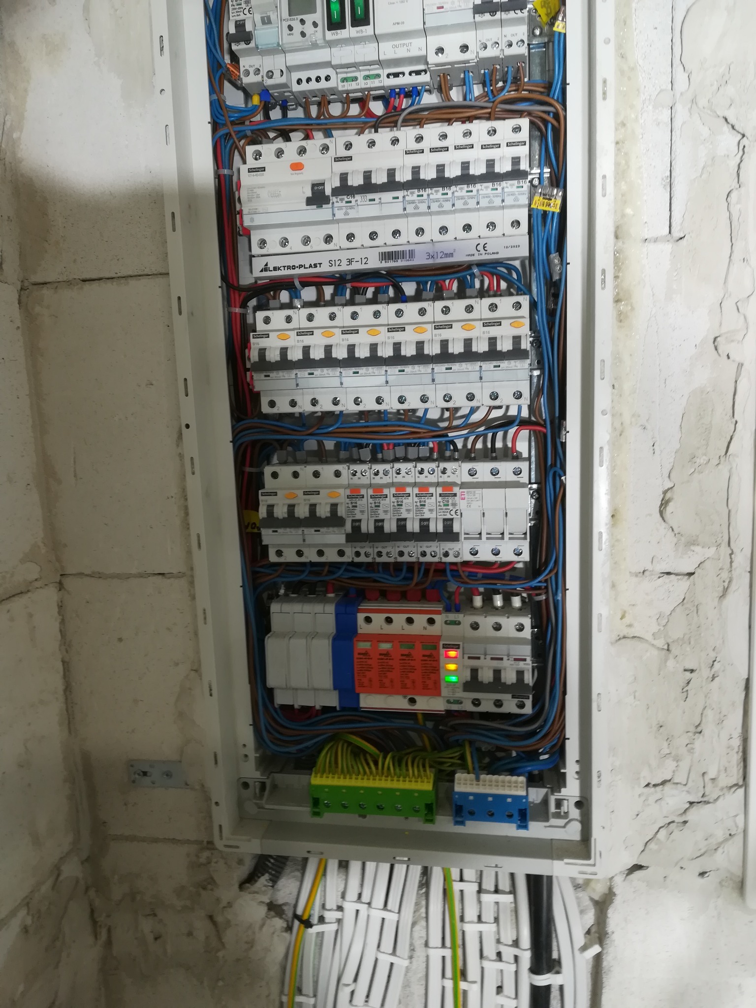 Otwarta skrzynka elektryczna z widocznymi bezpiecznikami, przewodami różnej grubości i koloru, zamontowana na surowej, otynkowanej ścianie. Widoczne elementy montażowe i kable w listwach...