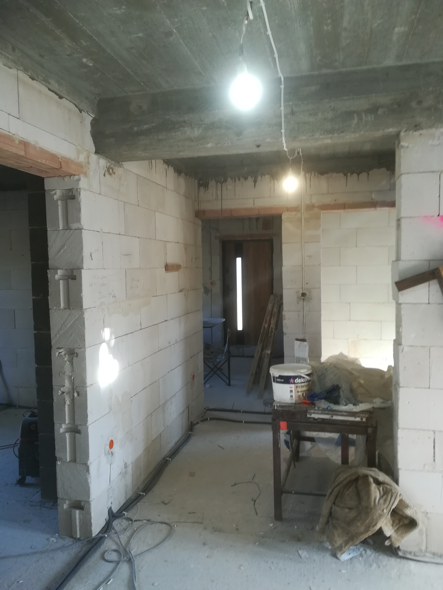 Wnętrze w stanie surowym: ściany z białego betonu komórkowego, widoczne przewody elektryczne prowadzone po podłodze i suficie, prowizoryczne oświetlenie żarówkami, narzędzia i materiały budowlane...