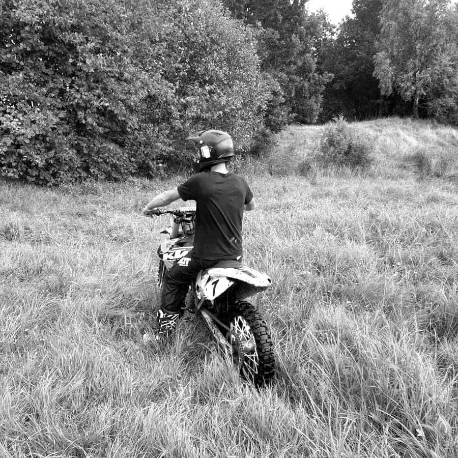 Czarno-białe zdjęcie motocyklisty w kasku siedzącego na motocyklu crossowym na łące z wysoką trawą, widok z tyłu.