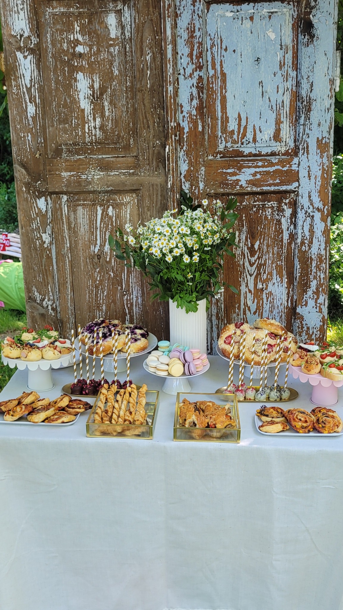 Stół z przekąskami i ciastami na białym obrusie, rustykalne drzwi w tle, bukiet białych kwiatów w prążkowanym wazonie, złote dekoracje i patery.