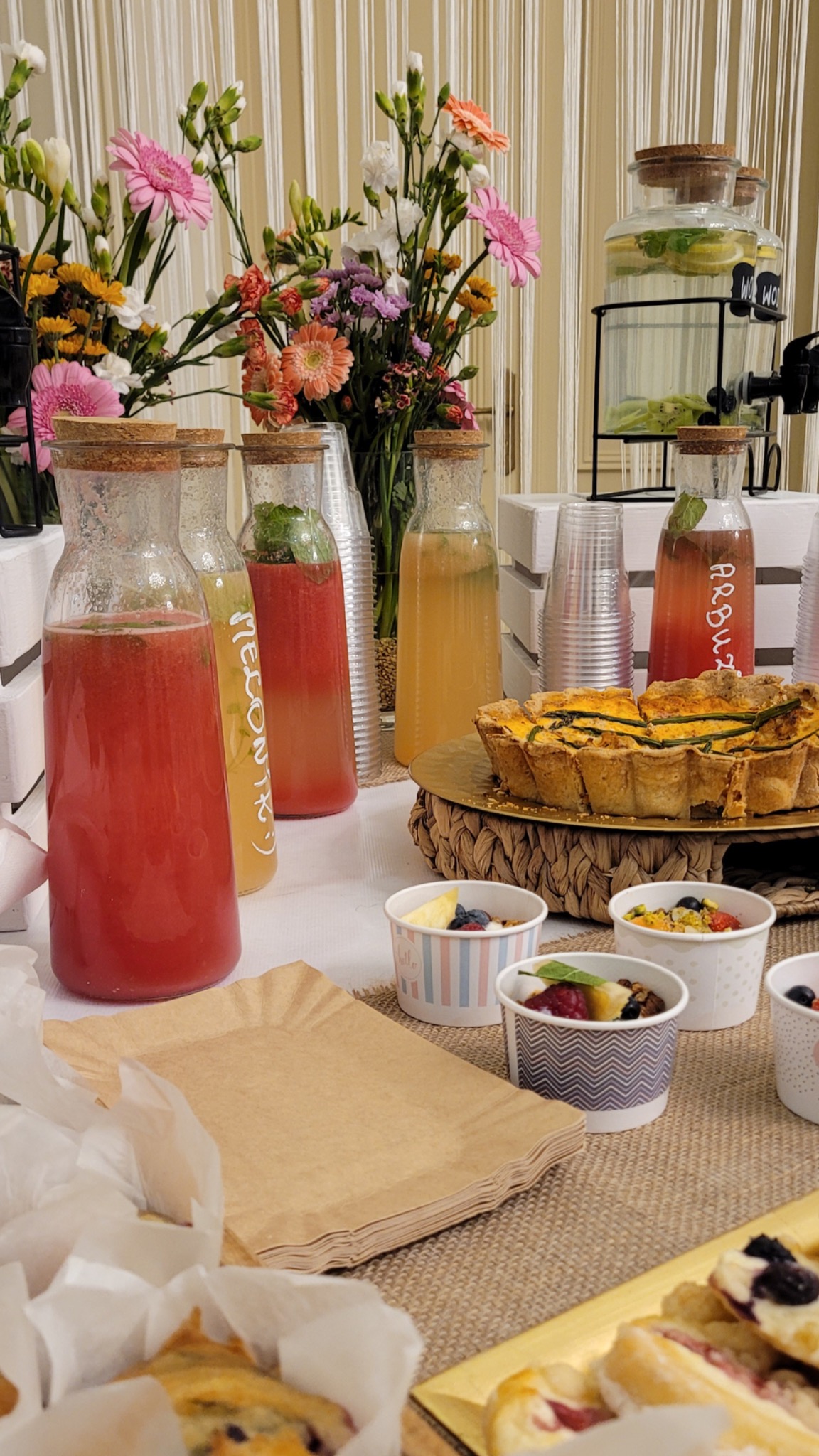 Stół cateringowy z dzbankami lemoniady (arbuzowa, melonowa) z korkowymi zamknięciami, quiche na plecionym podkładzie, deserami w papierowych kubeczkach, kwiatami w wazonach na tle białych...