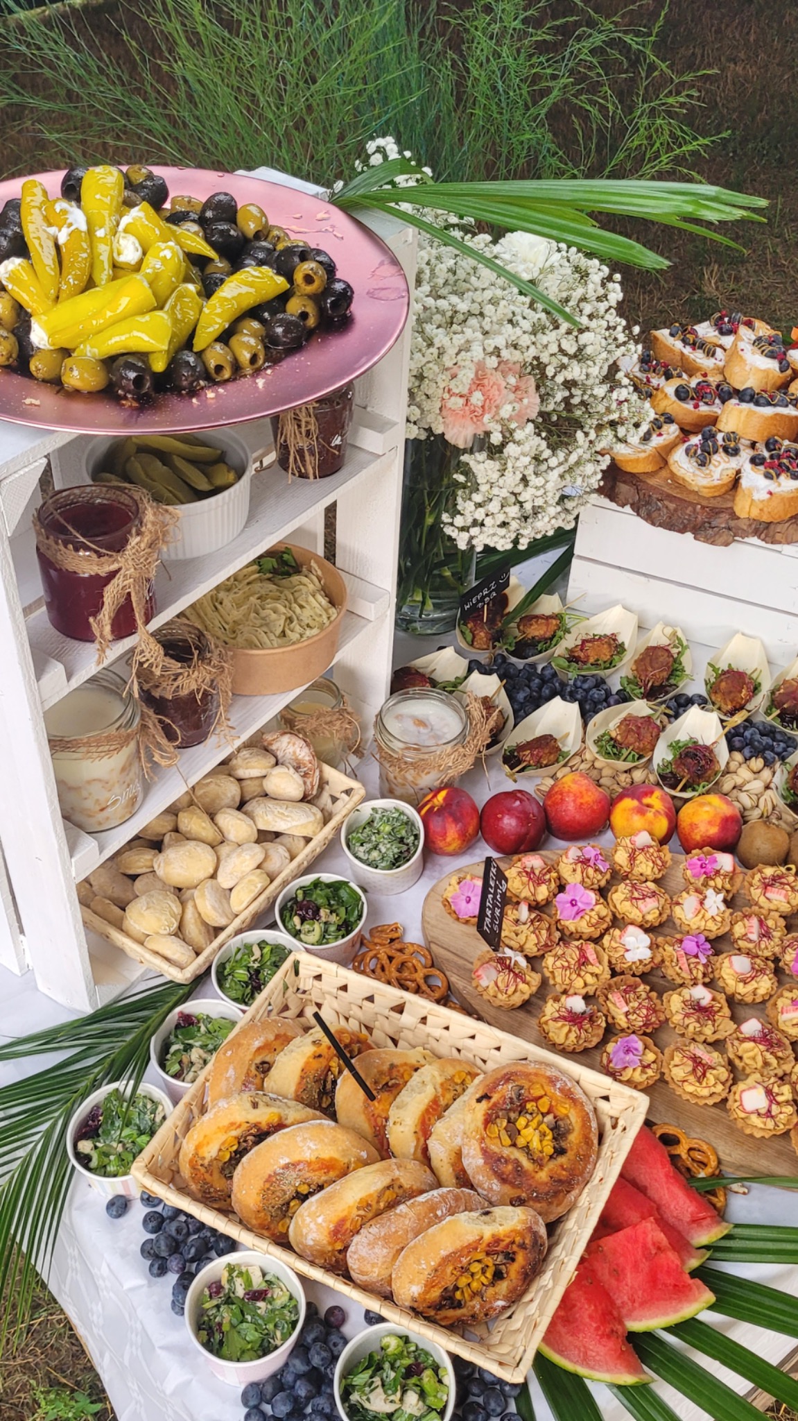 Bogato zastawiony stół z różnorodnymi przekąskami: oliwki z papryką, pieczywo, sałatki, tartaletki, owoce, słoiki z przetworami, ułożone na białym stole i drewnianych półkach na tle zieleni.