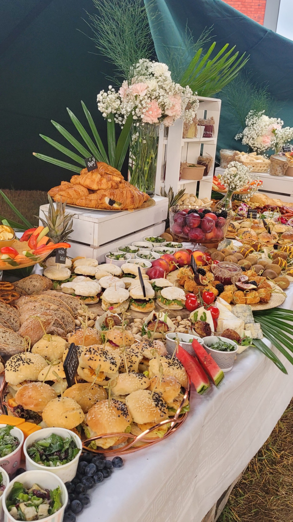 Bogato zastawiony stół z różnorodnymi przekąskami: mini burgery, kanapki, croissanty, owoce, sery i sałatki, udekorowany kwiatami i liśćmi palmowymi.