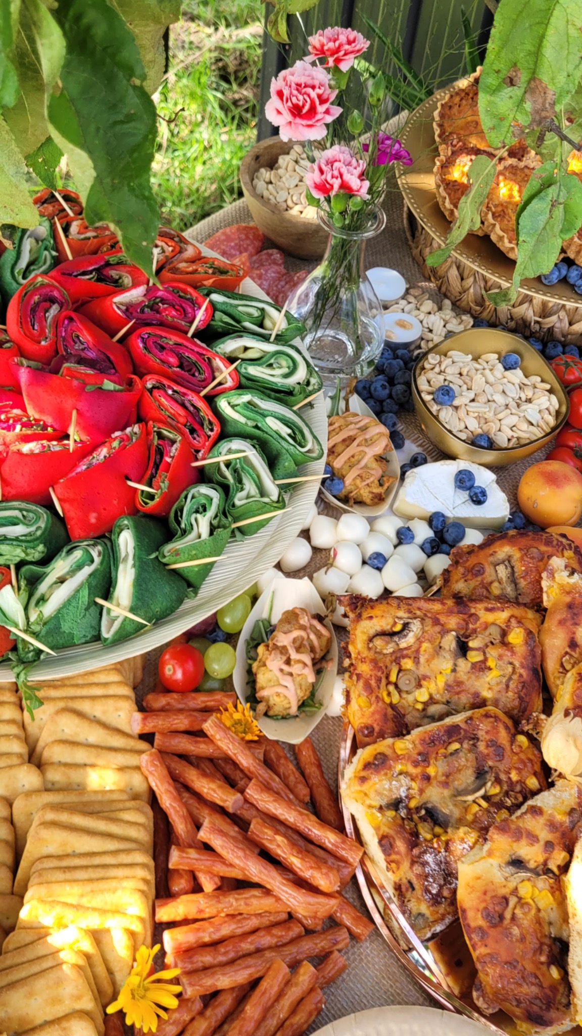 Bogato zastawiony stół z różnorodnymi przekąskami: kolorowe roladki na szpilkach, sery, wędliny, krakersy, pieczywo z grzybami i kukurydzą, orzechy, owoce, małe kulki mozzarelli i kwiaty w wazonie,...
