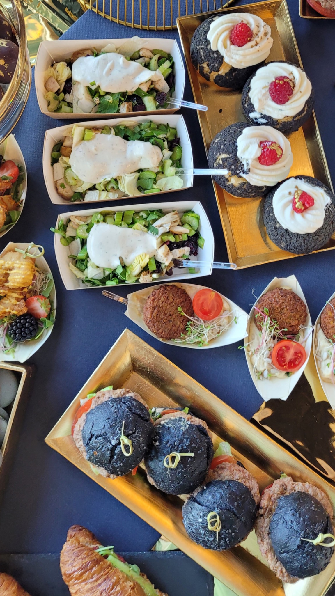 Ekskluzywny catering z mini burgerami z czarną bułką, sałatkami w papierowych pojemnikach, deserami z bitą śmietaną i owocami, podane na złotych tacach na ciemnym tle.