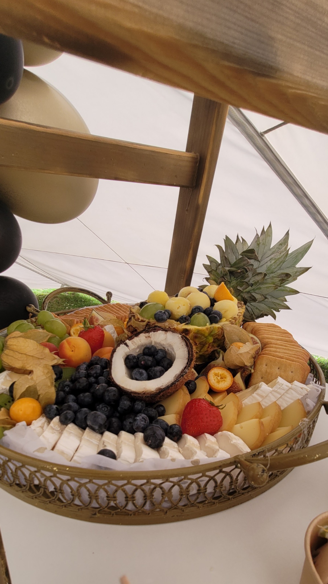 Dekoracyjna taca z owocami i serami: przekrój kokosa wypełniony borówkami, ananas z owocami, truskawki, brie i inne sery, krakersy, physalis, morele i winogrona, całość pod drewnianą konstrukcją...