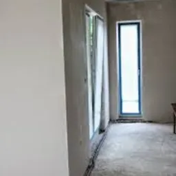 Wnętrze w trakcie prac wykończeniowych: świeżo nałożona gładź na ścianach, widoczne kable wzdłuż podłogi, okno z zasłoną i wysokie drzwi w niebieskiej ramie.